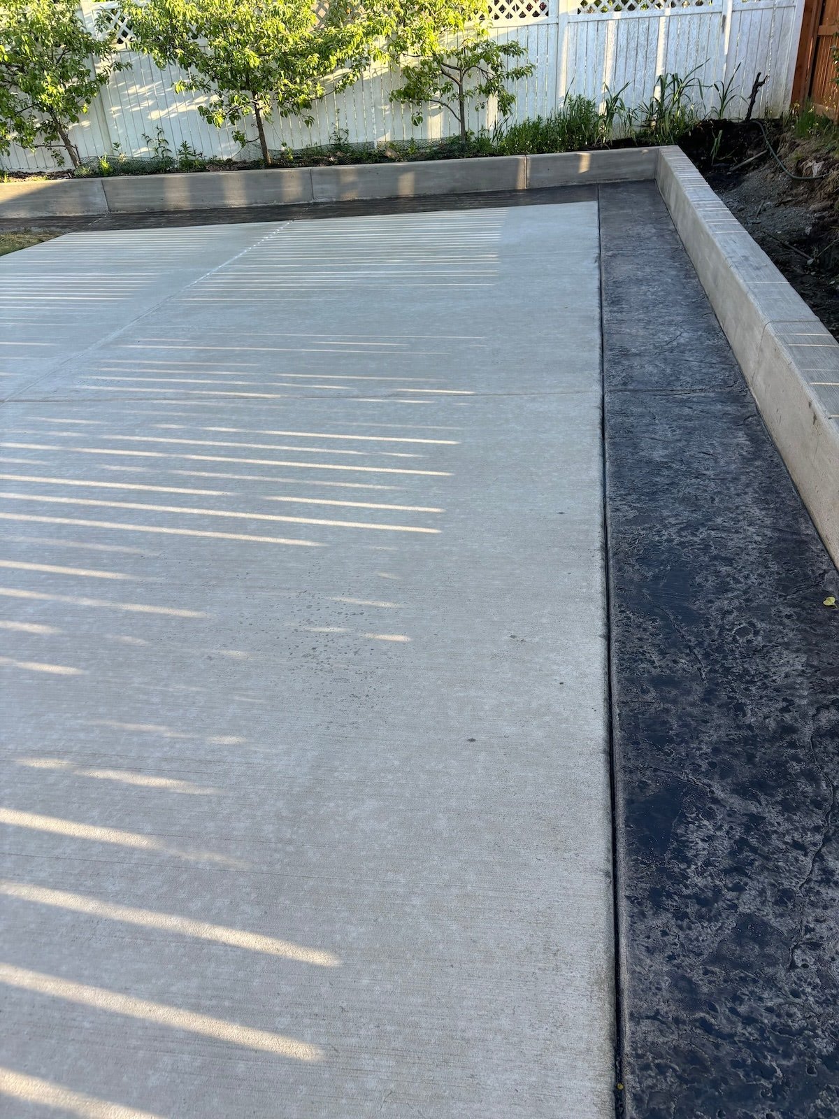 Concrete driveway with shadow patterns and bordered by a low retaining wall, with garden and white fence in the background.