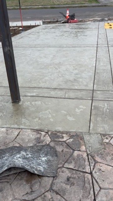 Freshly poured concrete driveway with adjacent textured concrete surface, traffic cone, and construction equipment in the background.