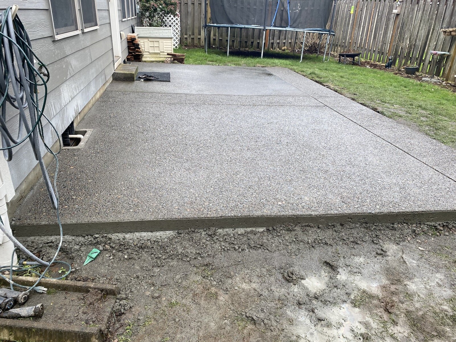 Freshly poured concrete patio in a backyard, adjacent to a house, with a garden hose on the side and a trampoline in the background.