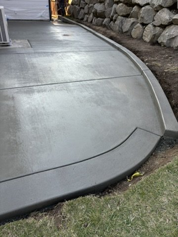 Freshly poured concrete patio with curved edges next to a stone wall and grass.