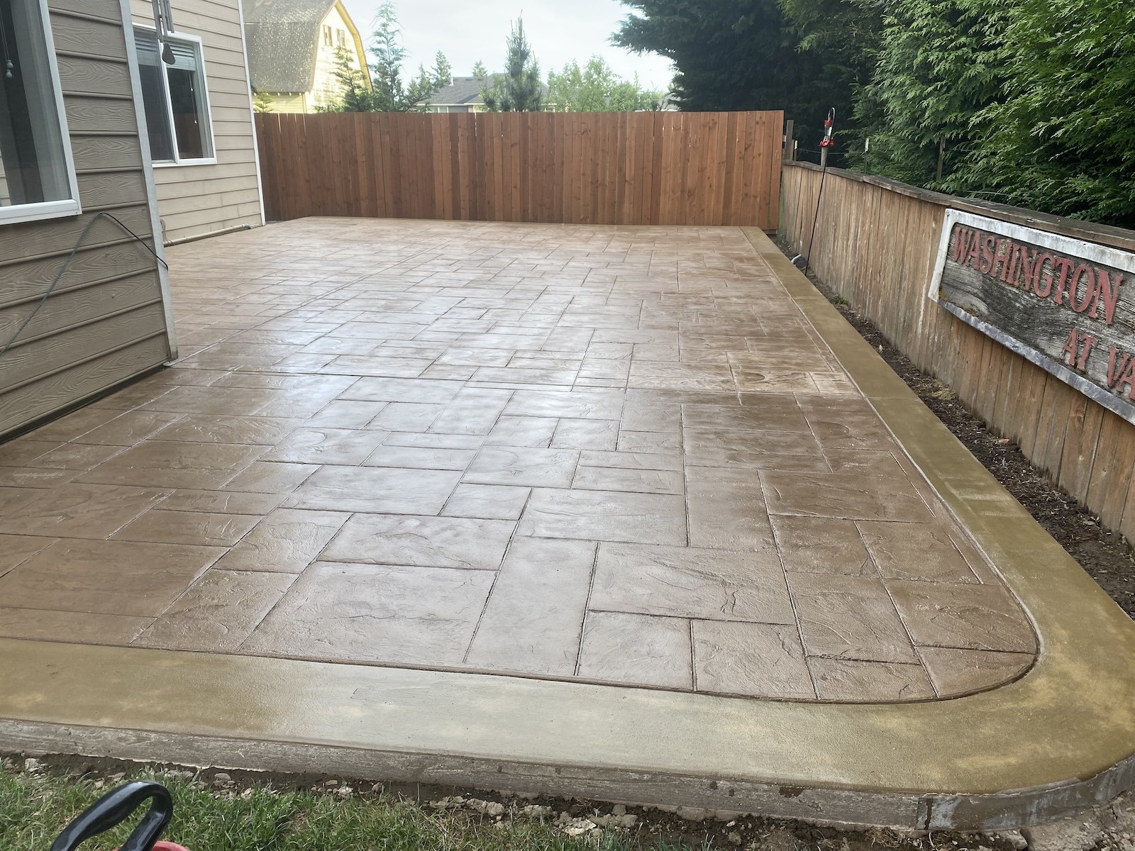 Freshly laid stamped concrete patio with a wooden fence and house siding in the background. There is a wooden sign on the right side of the fence.