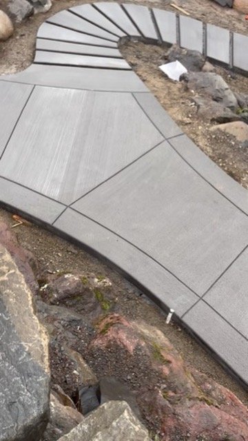 Curved concrete pathway under construction with geometric patterns, surrounded by rocks and soil.