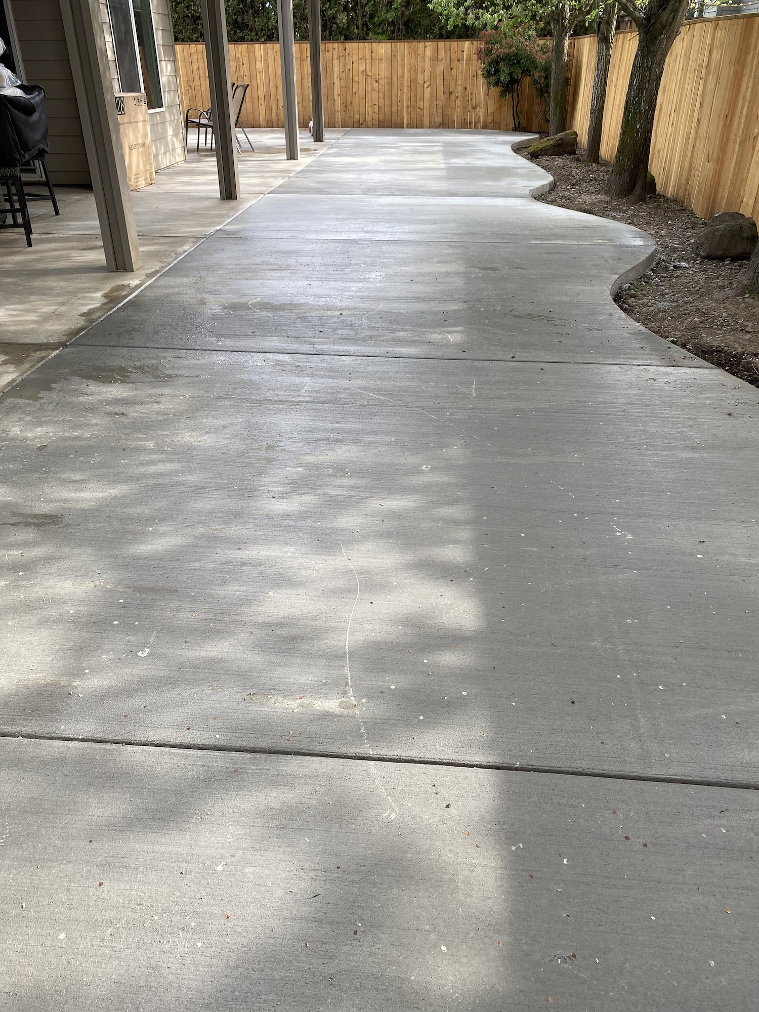 Concrete patio with curved edges, adjacent to a wooden fence and trees, providing shaded areas.