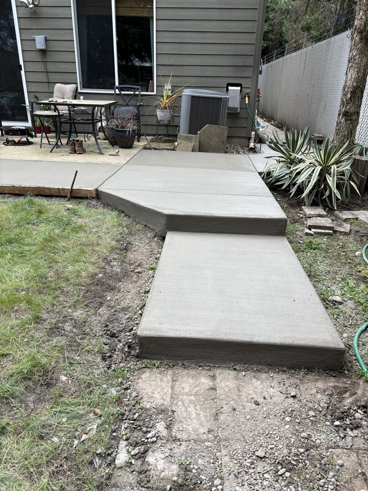 Freshly poured concrete path leading to a patio with outdoor furniture, next to a house and surrounded by grass and plants.