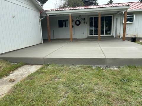 Freshly poured concrete patio with wooden posts and sliding glass doors, attached to a white house with a red metal roof, surrounded by grass.