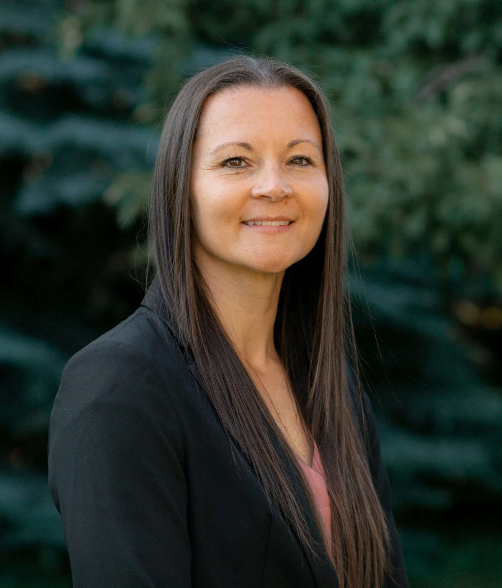 Woman with long brown hair smiling at the camera, wearing a black jacket, with green foliage in the background.