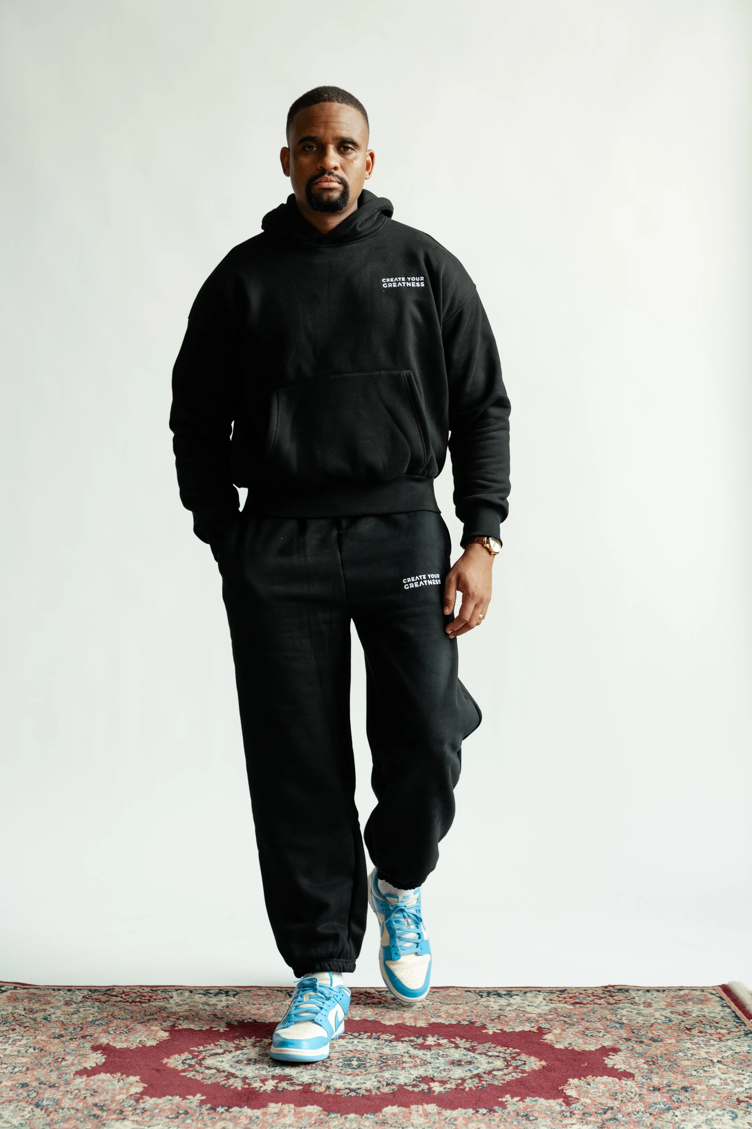 A man standing on a patterned rug, wearing a black hoodie and sweatpants with 'CREATE YOUR GREATNESS' printed on them, paired with blue and white sneakers.
