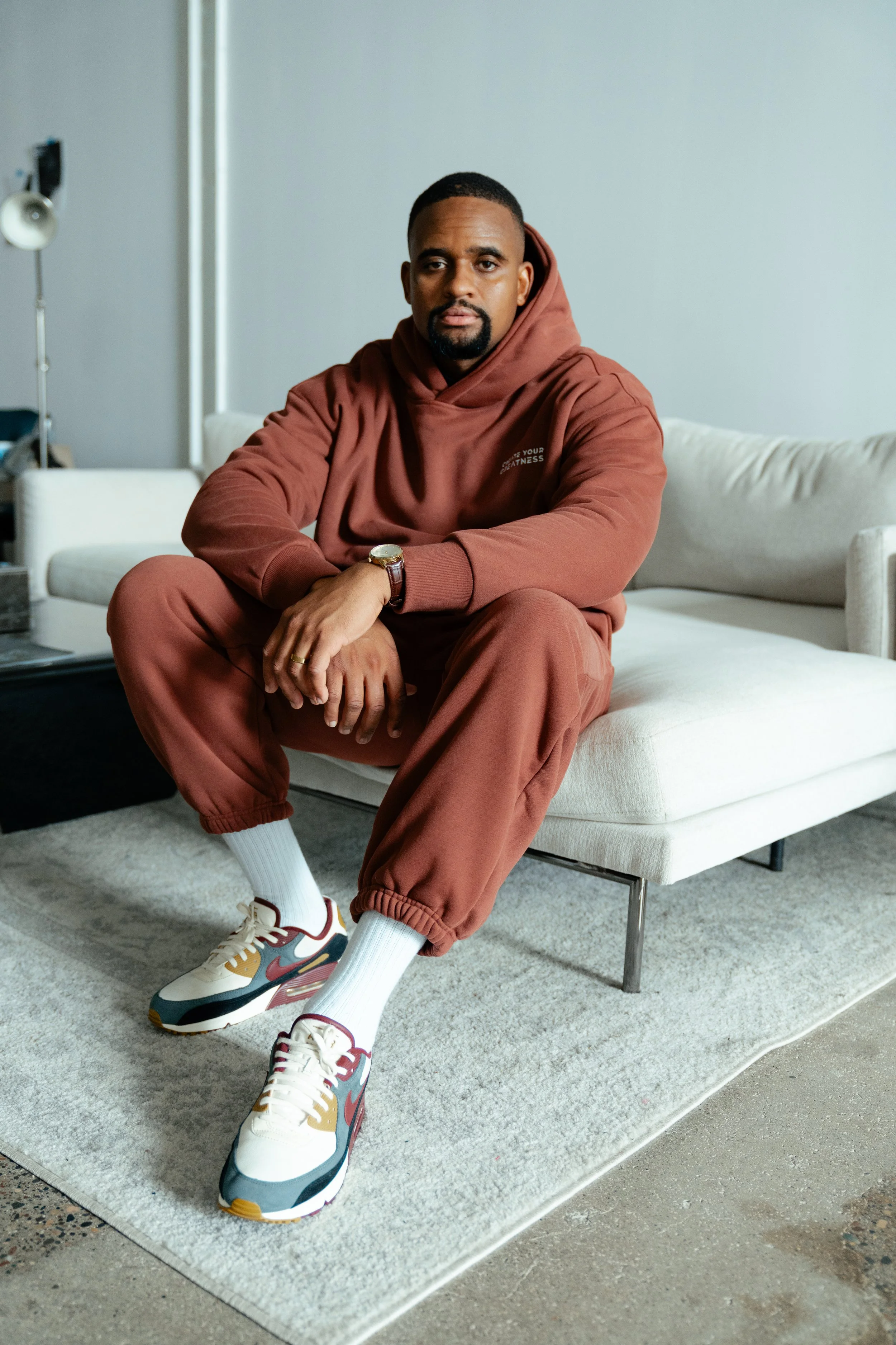A man sitting on a white couch in a room, wearing a brown hoodie and matching brown sweatpants, white socks, and white sneakers with multicolored accents.