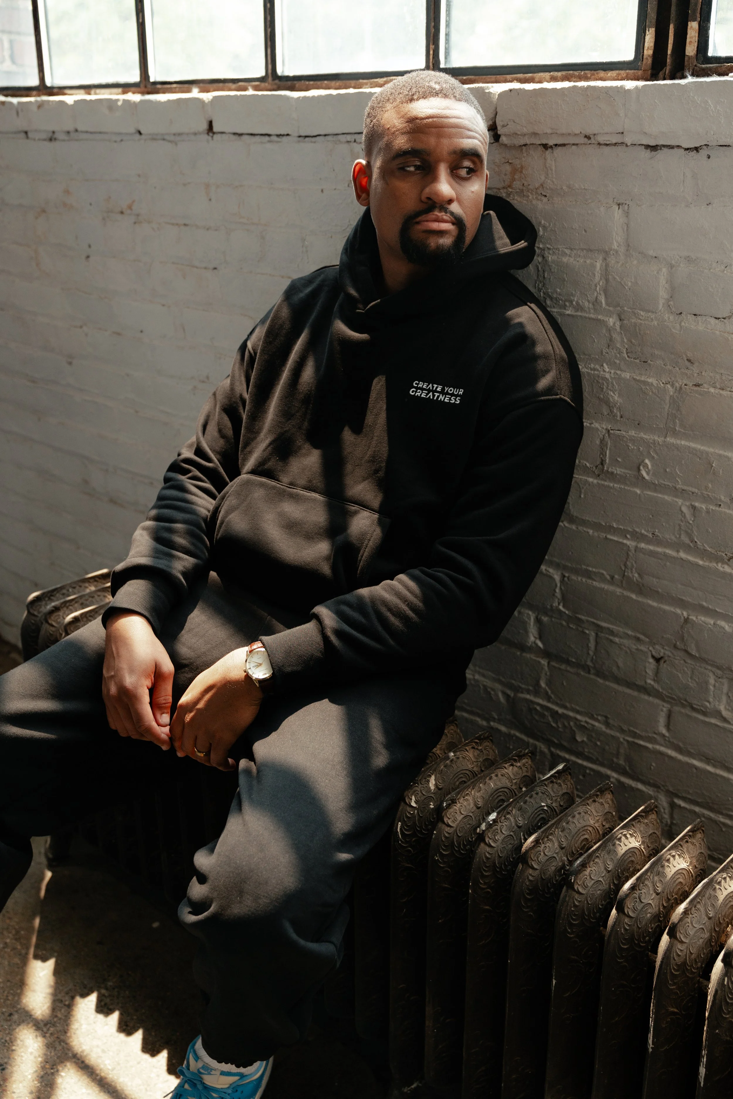 Man sitting on a vintage radiator near a white brick wall with large windows, wearing a black hoodie, gray pants, and sneakers, looking thoughtfully to the side.