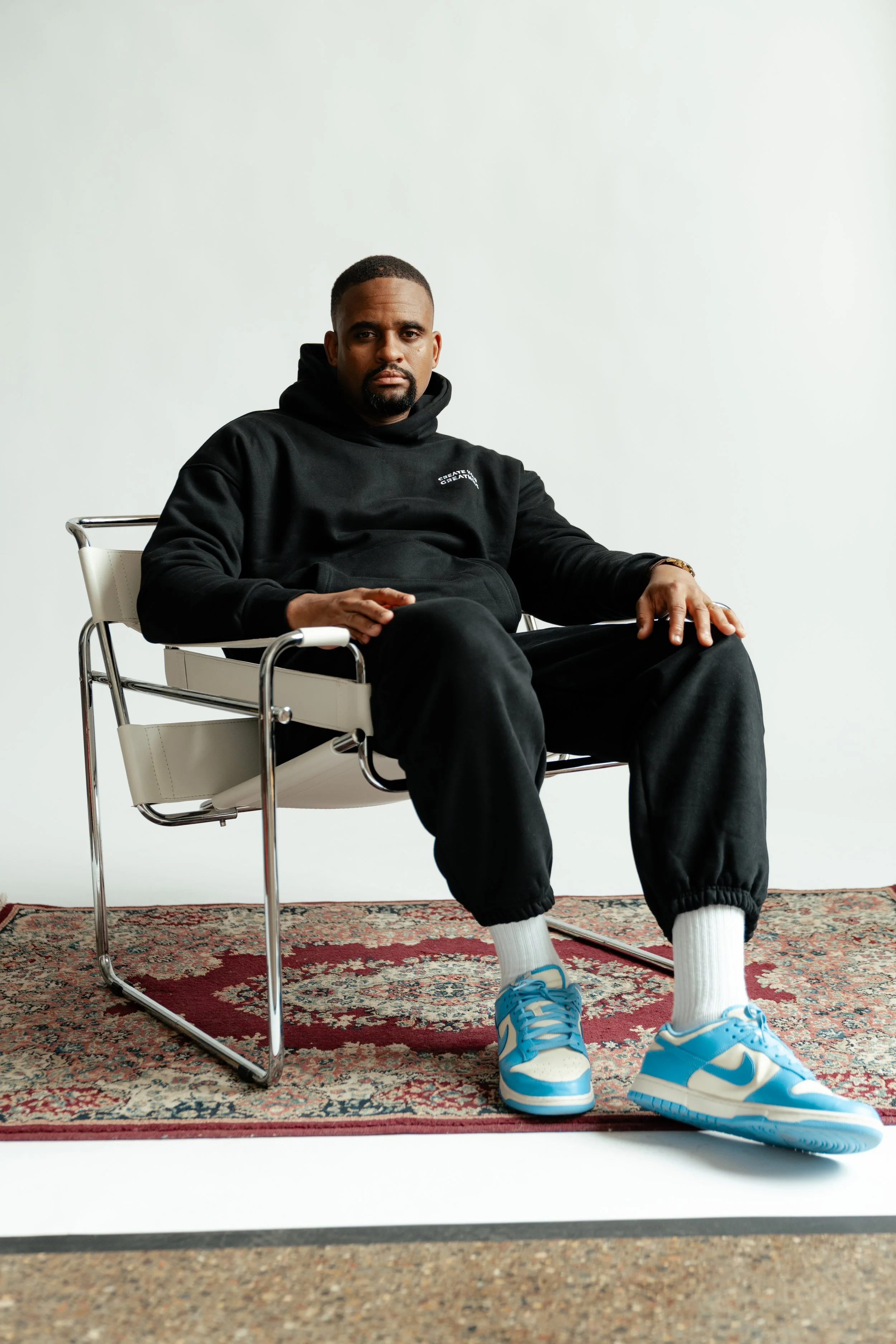 Man sitting on a modern white and chrome chair on a patterned rug, wearing a black hoodie, black joggers, white socks, and blue and white sneakers.