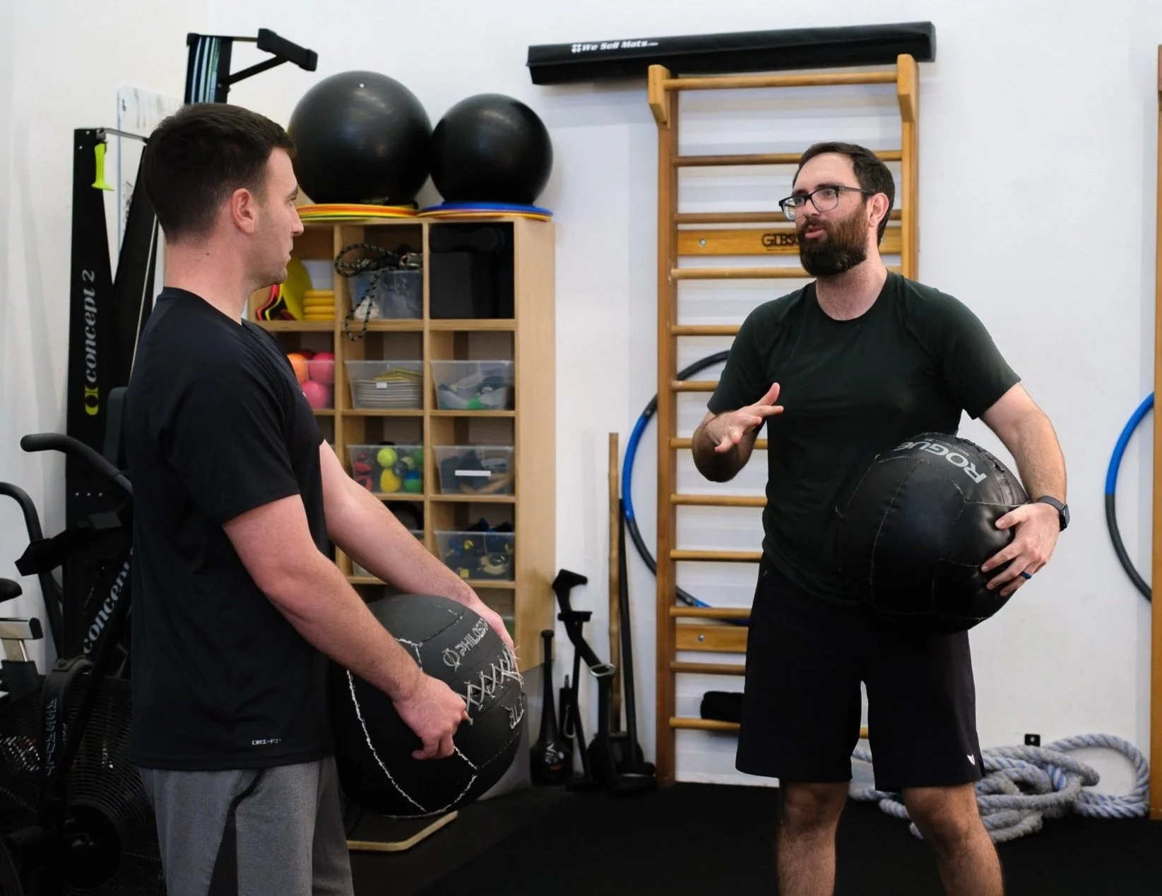 Two men in a gym, one holding a black medicine ball and the other gesturing while speaking, with gym equipment like large balls, a wooden wall ladder, and resistance bands in the background.