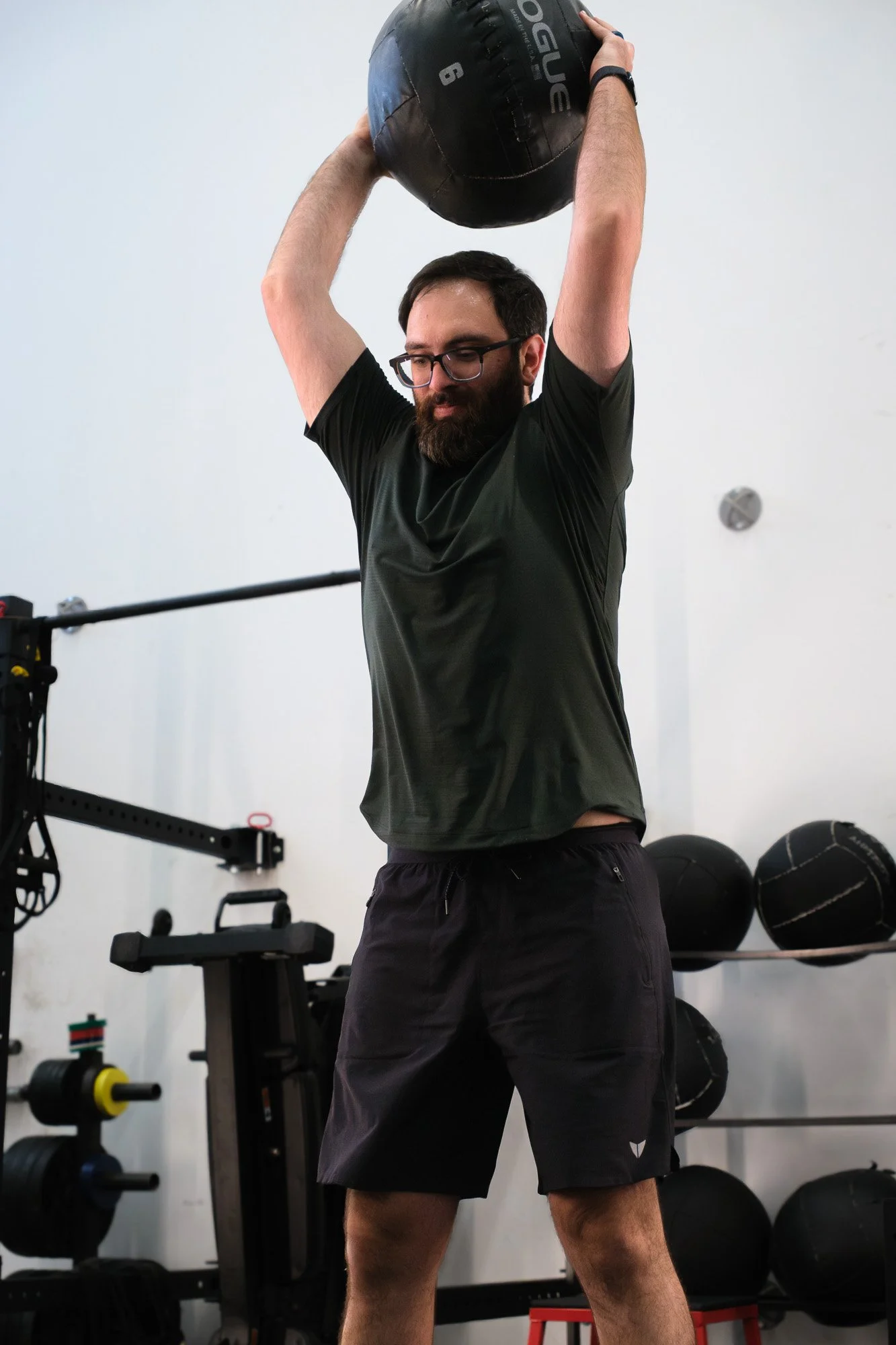 A bearded man with glasses lifting a black medicine ball overhead in a gym.