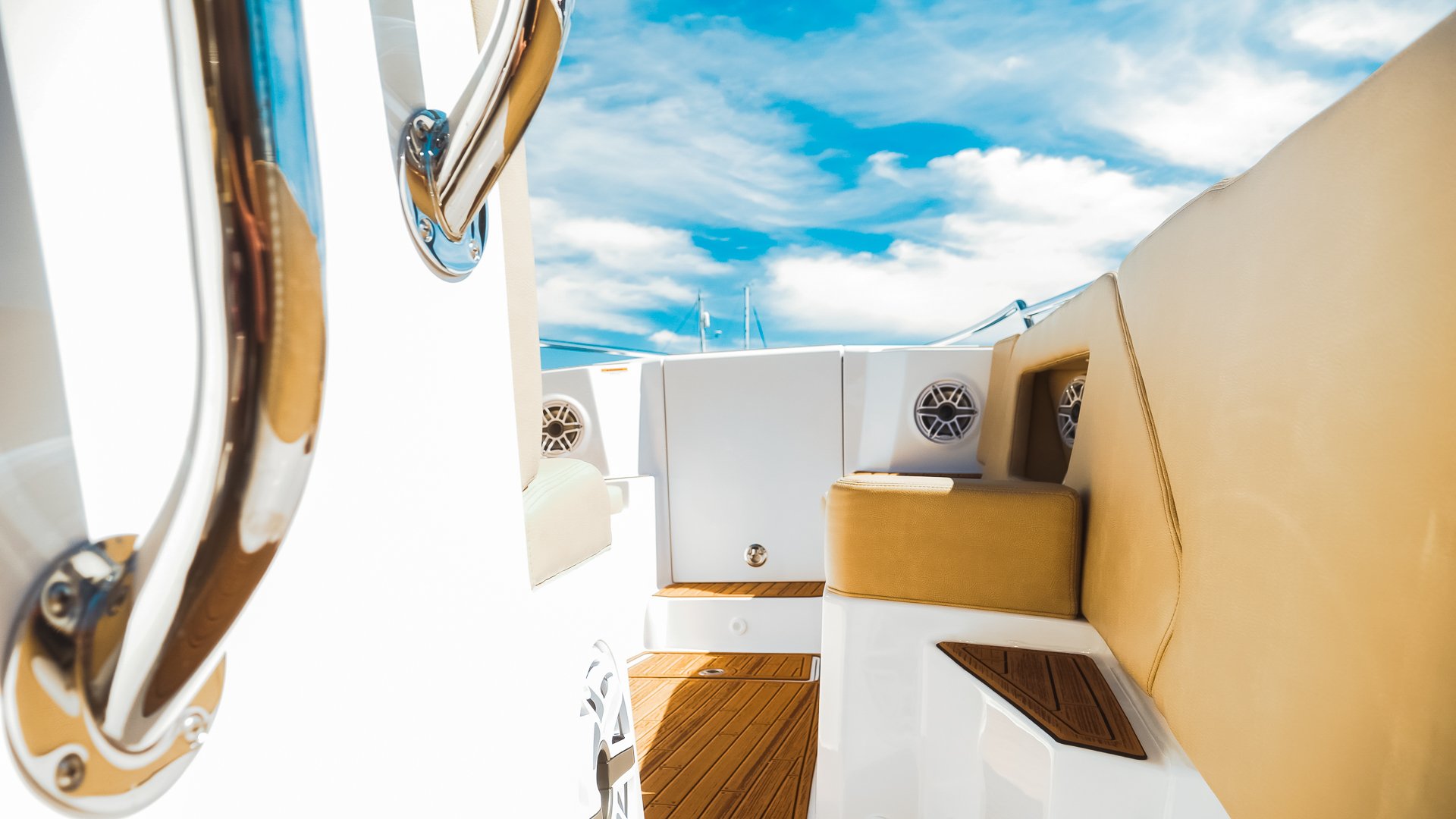 View of a boat deck with beige cushioned seats, wooden flooring, and a chrome railing, under a partly cloudy sky.