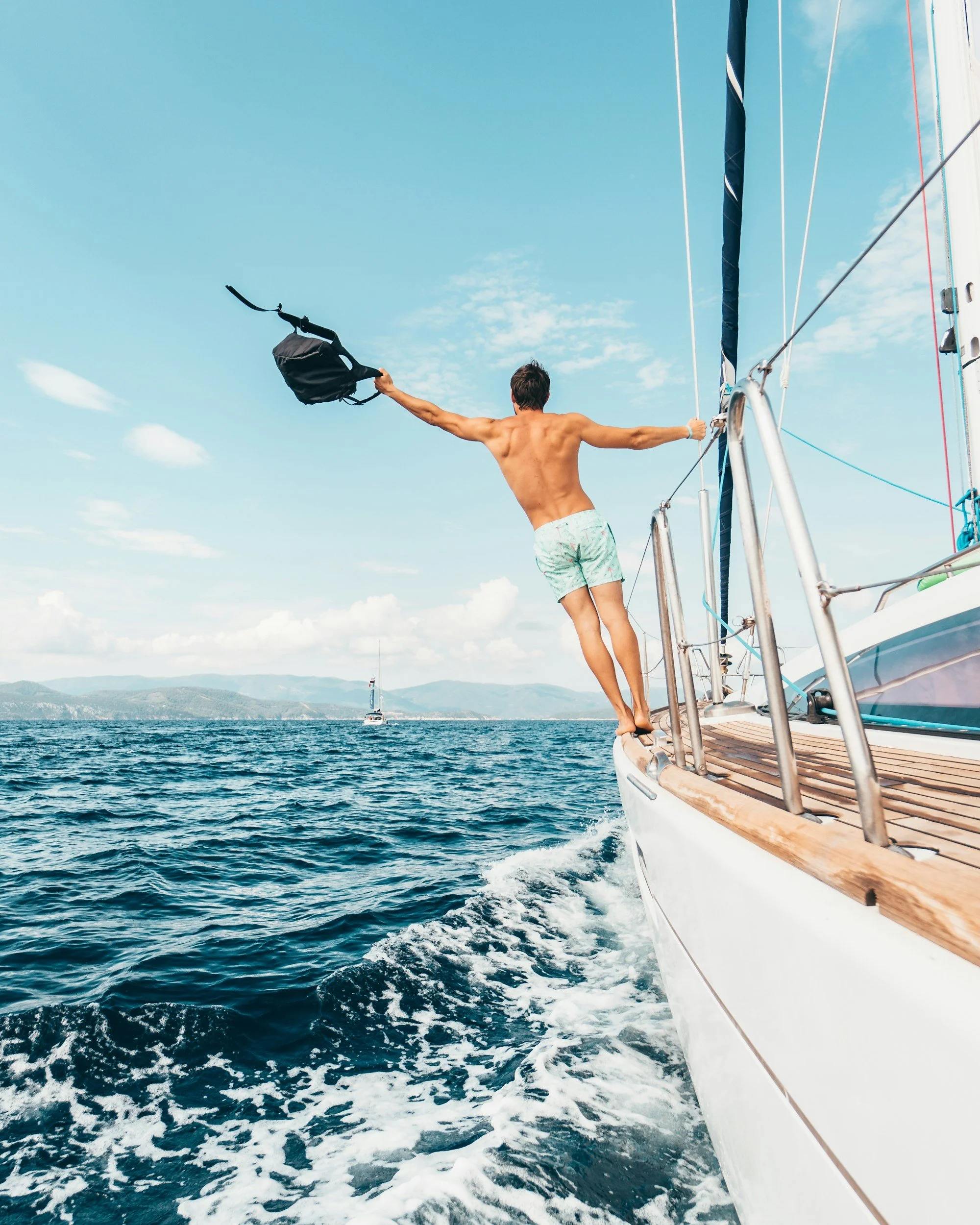 A man in swim trunks standing on the edge of a boat with arms outstretched, holding a bag in one hand, on a body of water with a sailboat in the background.