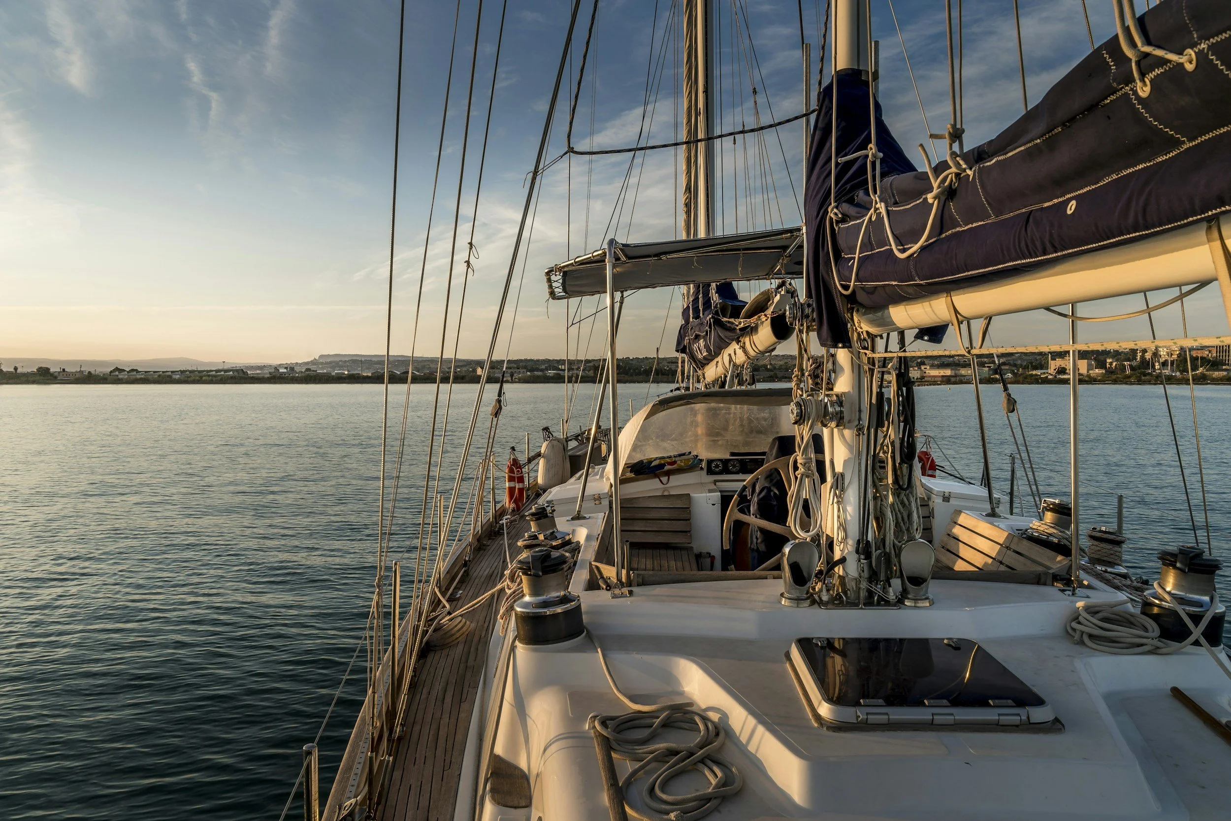 A sailboat on calm water during sunset with distant shoreline and buildings in the background.