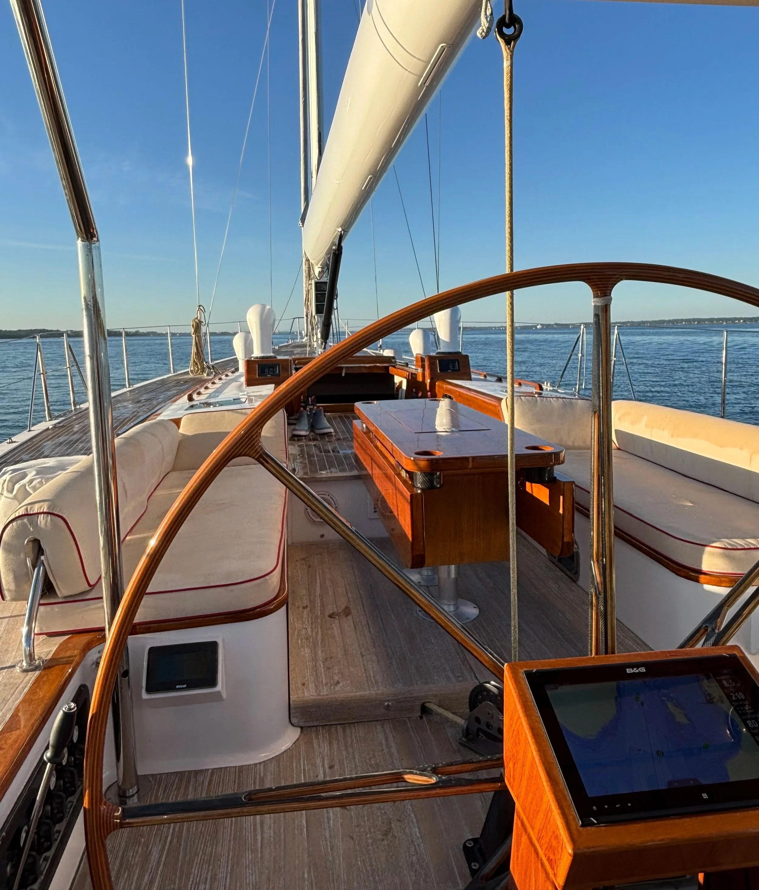 View of the deck and cockpit of a sailing yacht with a large steering wheel, navigation equipment, and seating area, on calm water during daytime with clear sky.