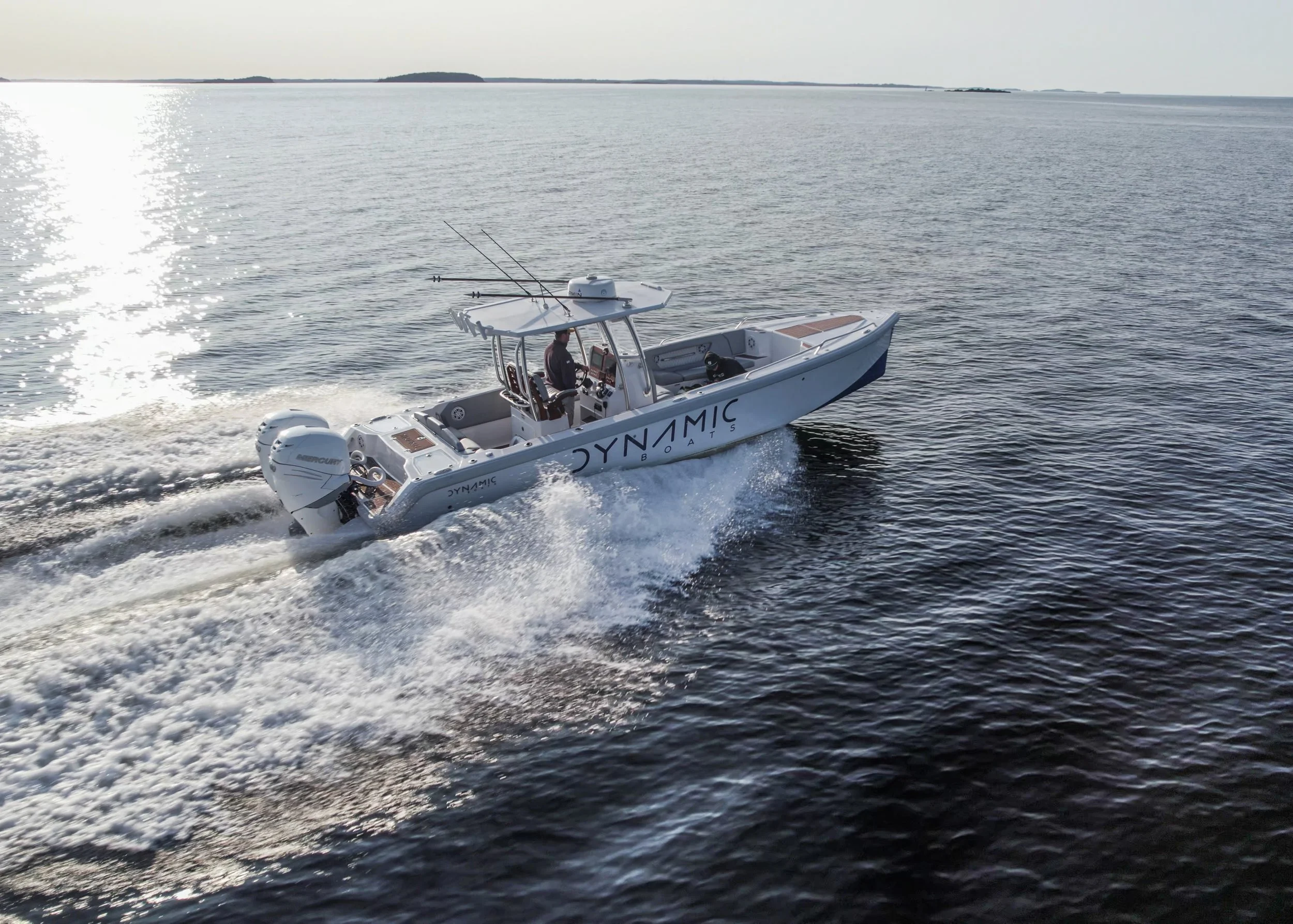 A white Dynamic Boats motorboat speeds across open water, creating a trail of white foam behind it with an island or landmass visible in the distance under a clear sky.
