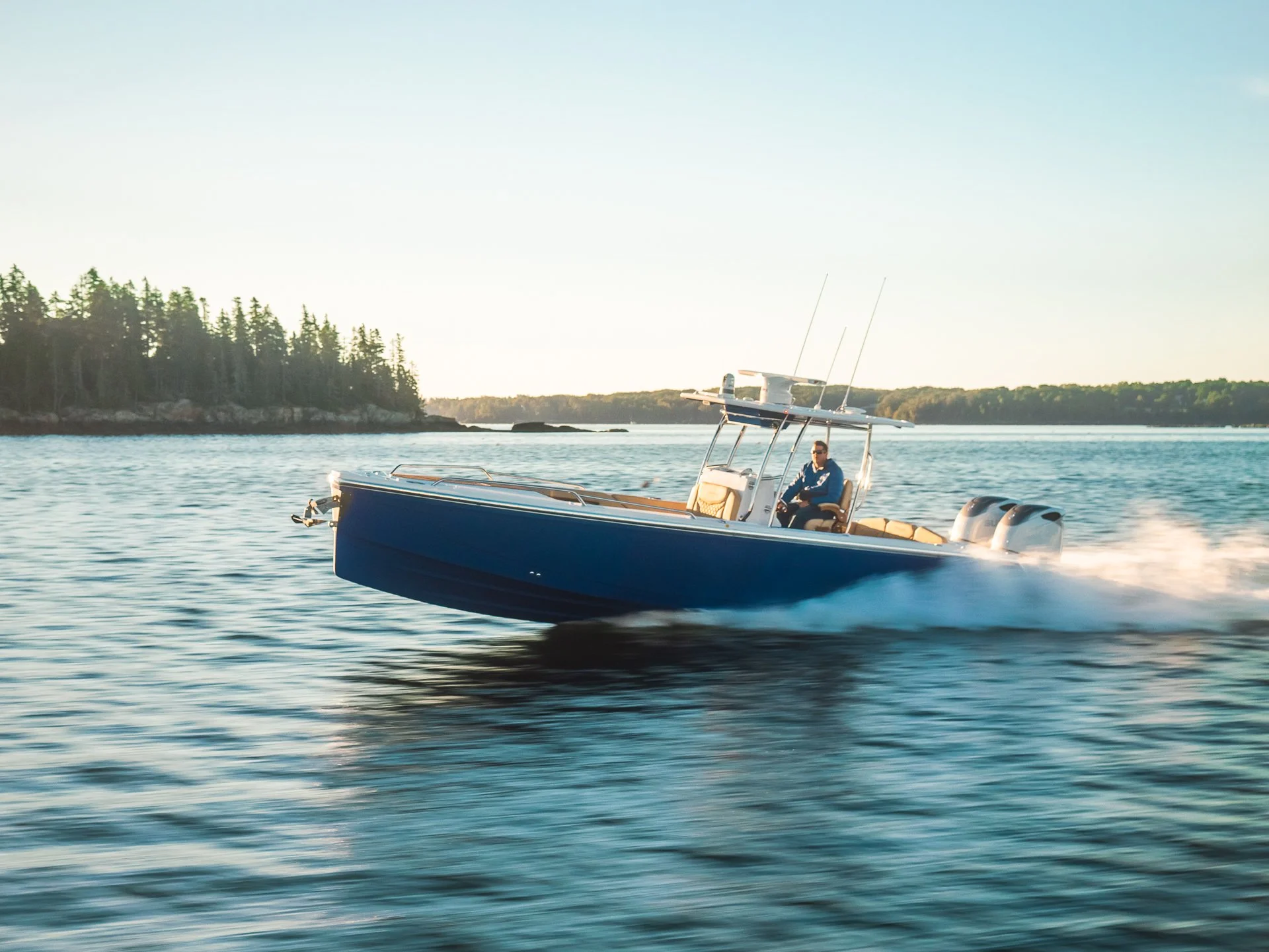 A blue speedboat with two outboard motors speeding across a body of water near a forested shoreline.