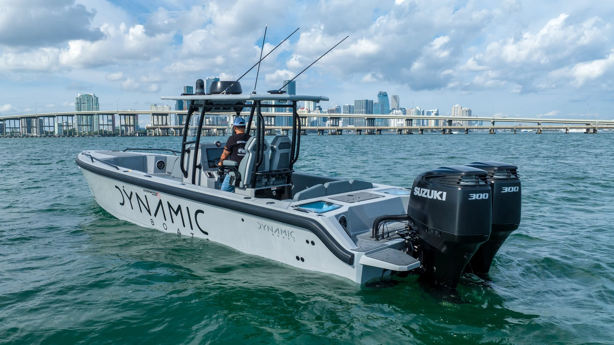 A boat with two Suzuki engines on the water, with a city skyline and bridge in the background.