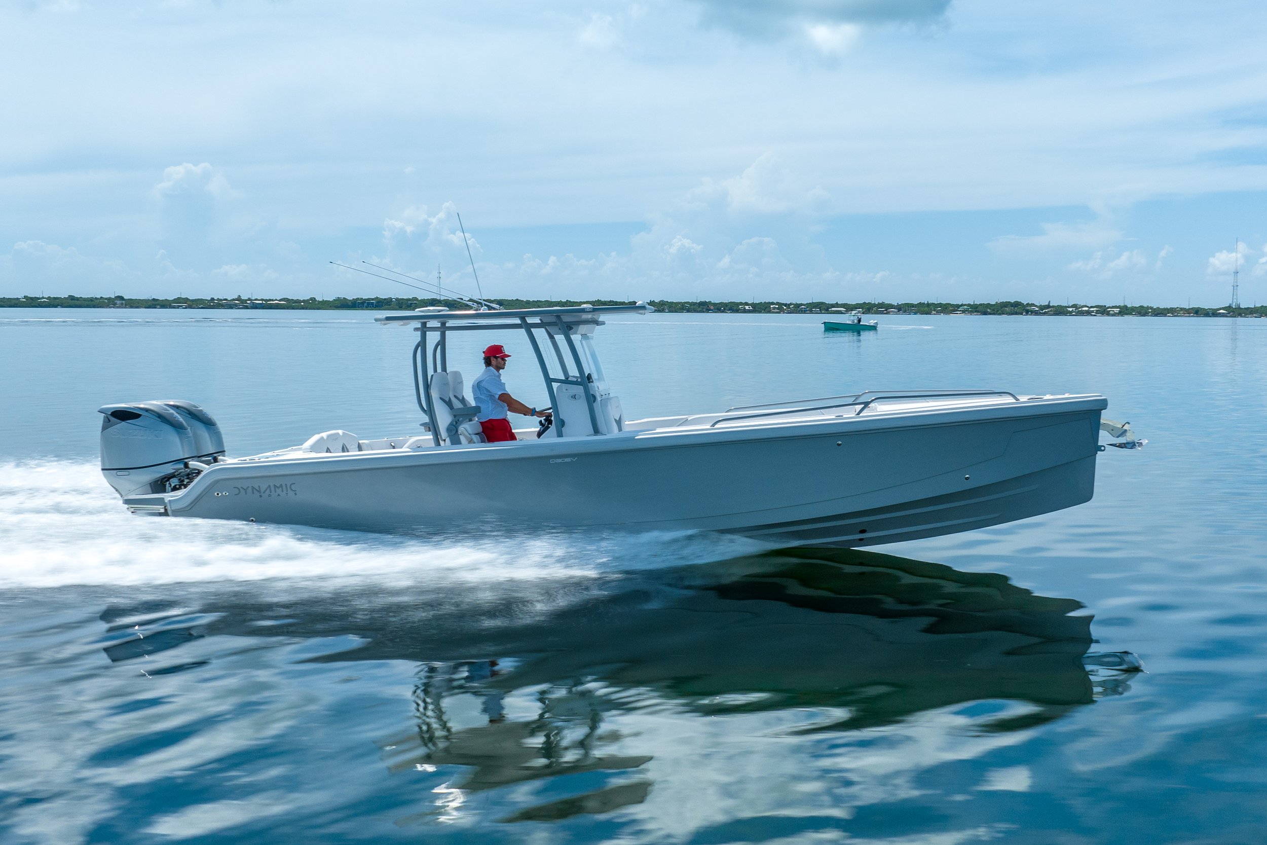 A person in a red cap and red shorts driving a sleek, white motorboat on a calm body of water. The boat has a canopy and is equipped with a powerful outboard engine, creating a wake as it moves. The background shows a distant shoreline with some stru