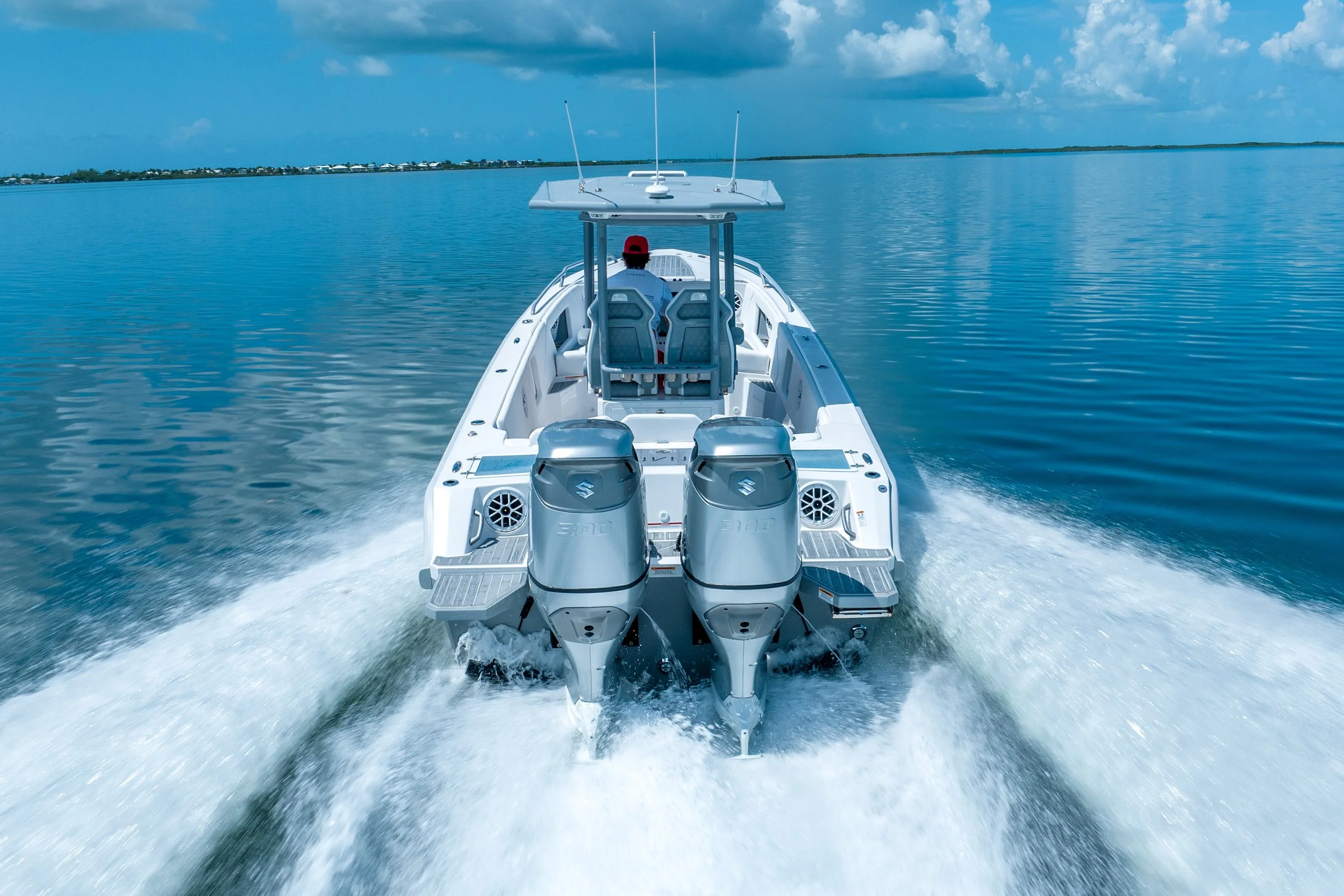 A boat with two engines cruising through calm blue water with a distant shoreline and partly cloudy sky in the background.