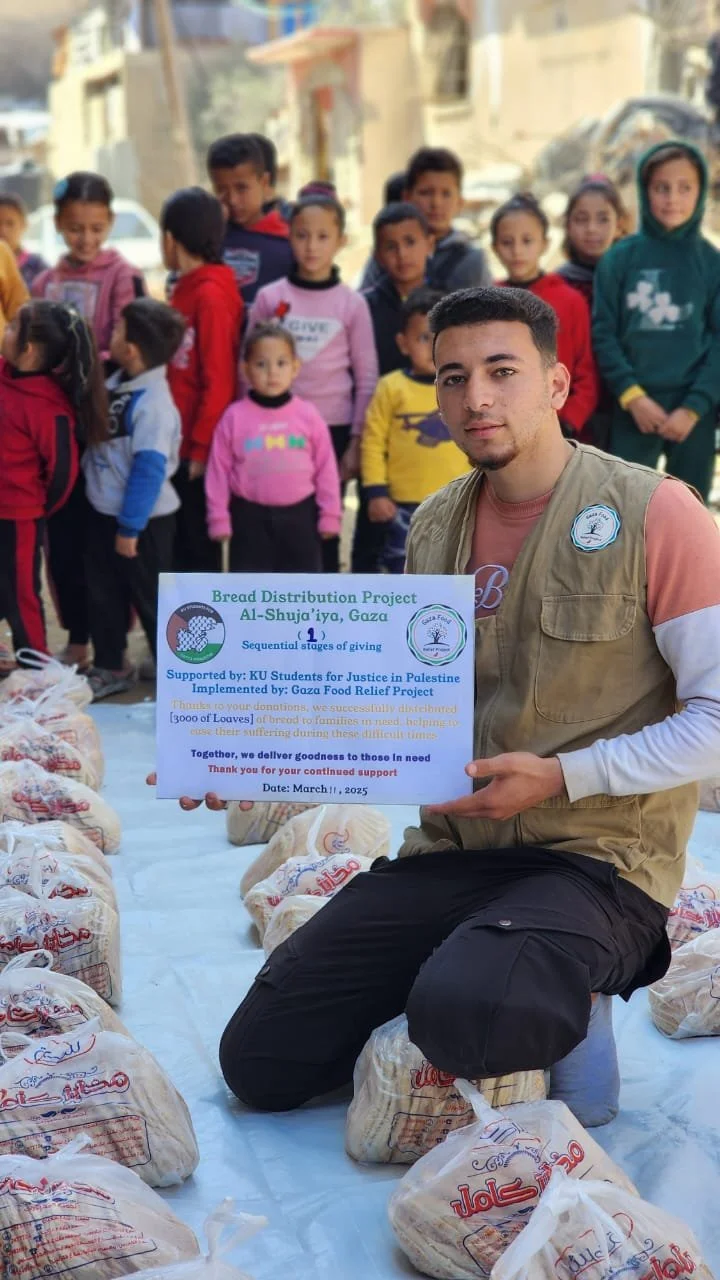 A young man kneeling, holding a sign for a bread distribution project in Al-Shuja'iya, Gaza, surrounded by bags of bread and children in the background. The sign mentions support from KU Students for Justice in Palestine and the Gaza Food Relief Project.