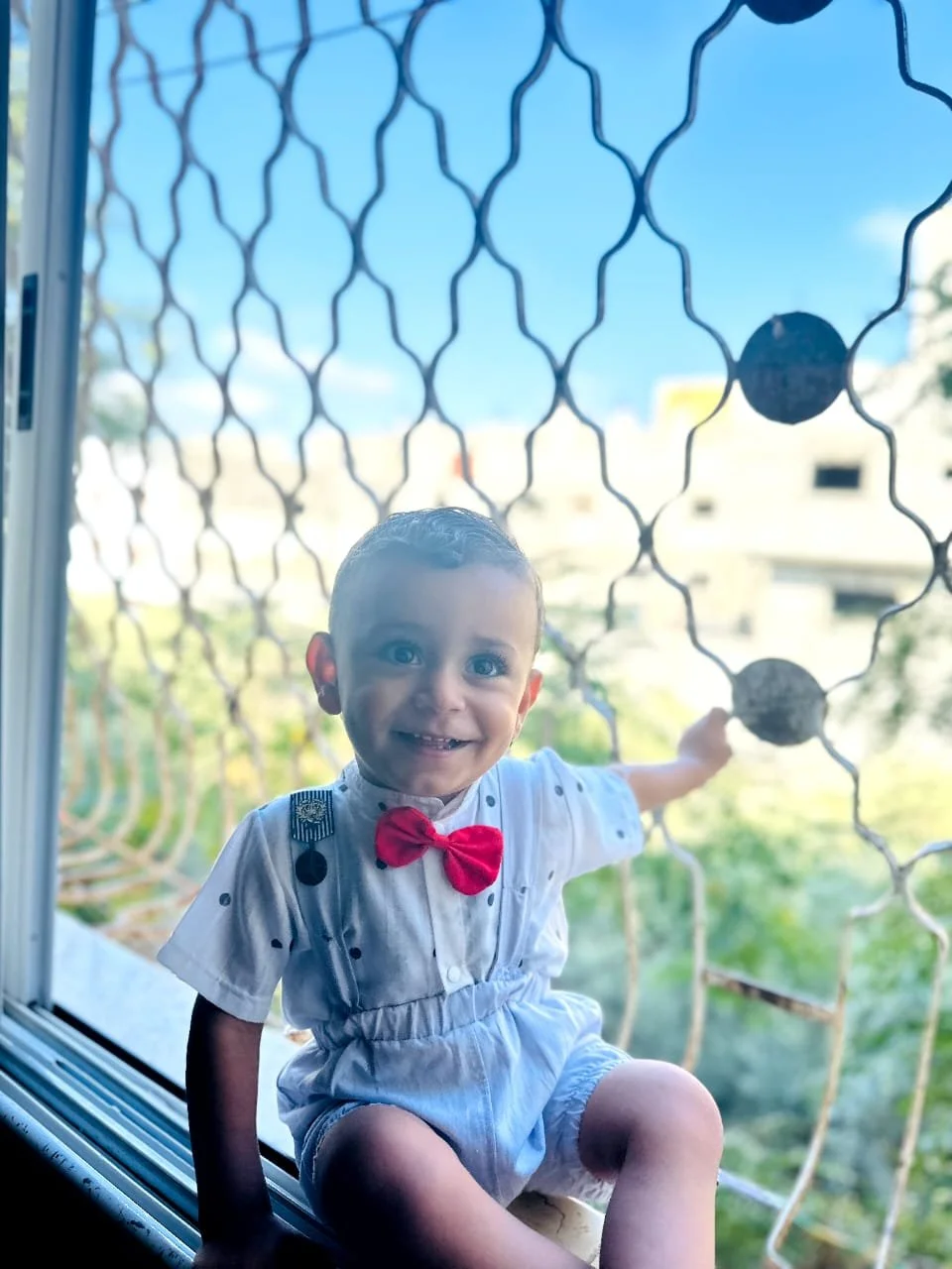 A smiling baby in a white outfit with a red bow tie sitting by a window with a patterned metal grill, with a blurred outdoor background.