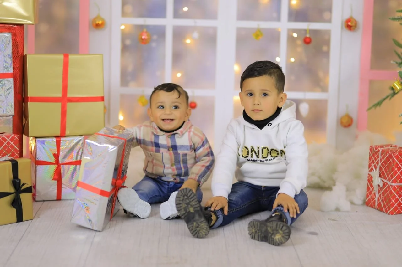 Two young children sitting in a festive setting with wrapped gifts, a decorated Christmas tree, and illuminated background.