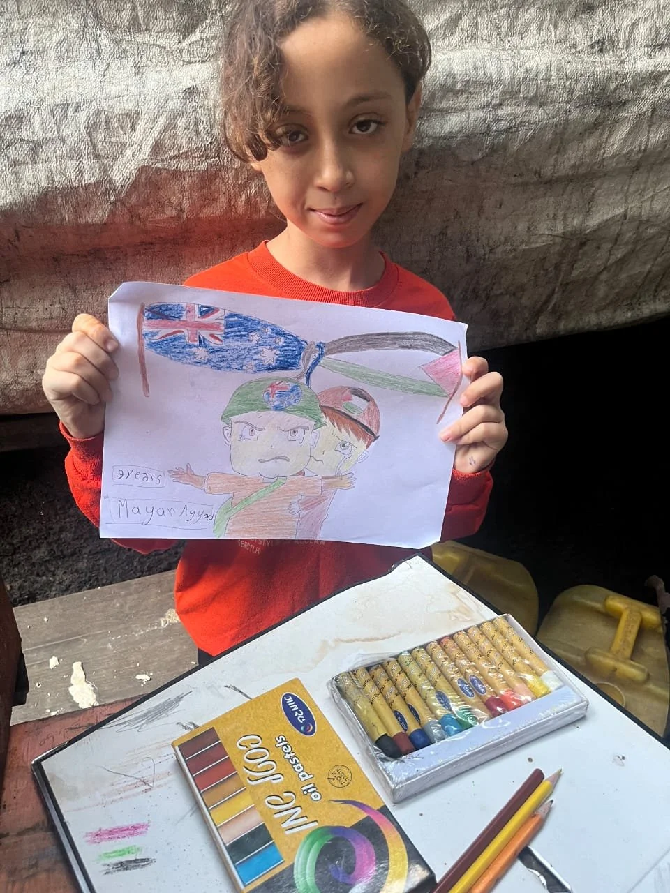 Child holding drawing with flags and characters, colored with oil pastels.