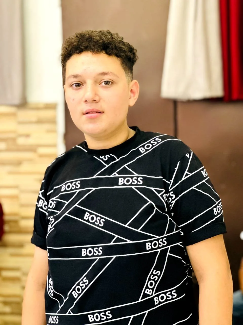 A young man with short curly hair wearing a black T-shirt with 'BOSS' print, standing indoors against a neutral background with curtains.