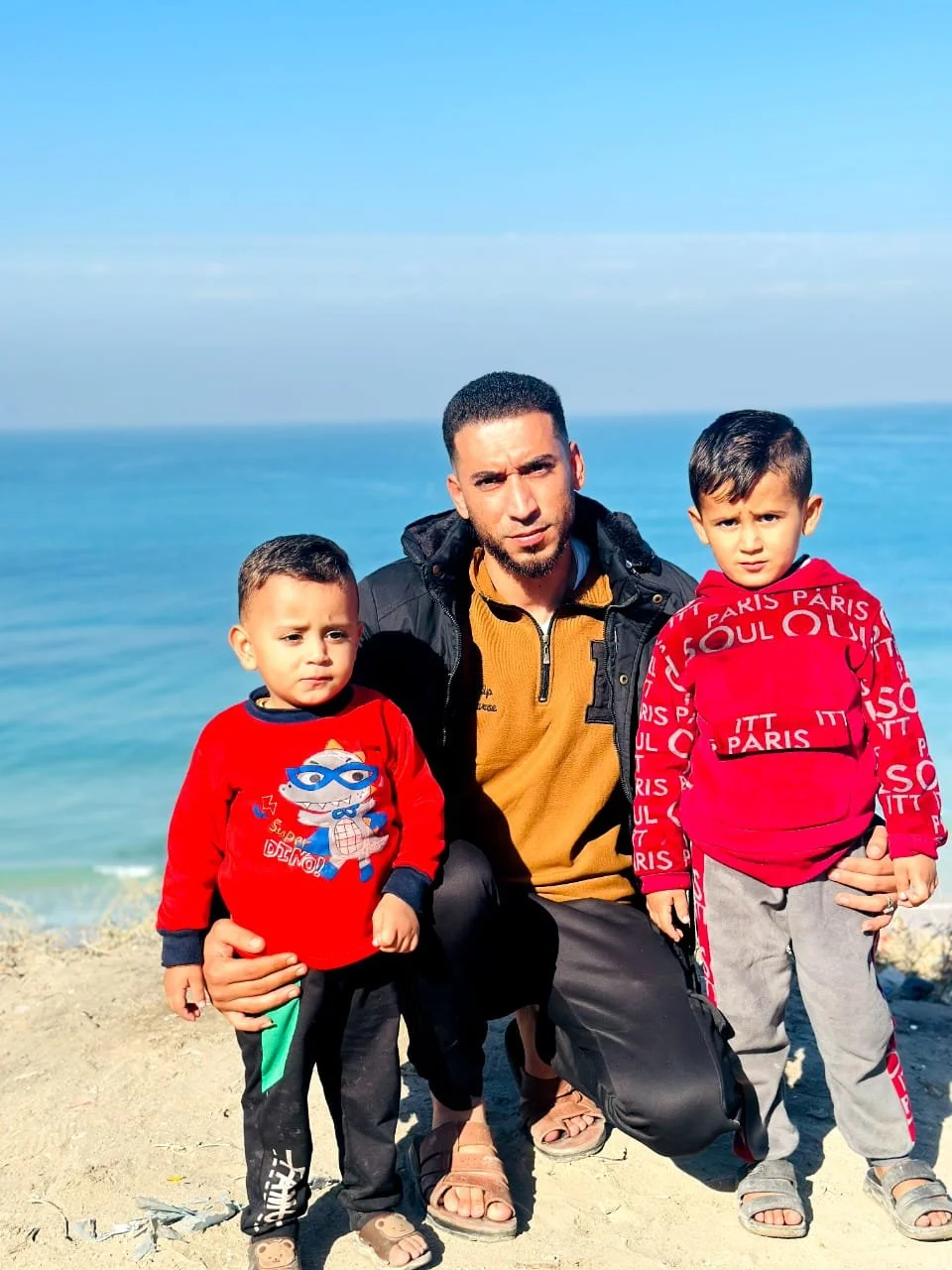 A man crouching on a sandy beach with two young boys, both wearing red sweaters, and the ocean visible in the background.