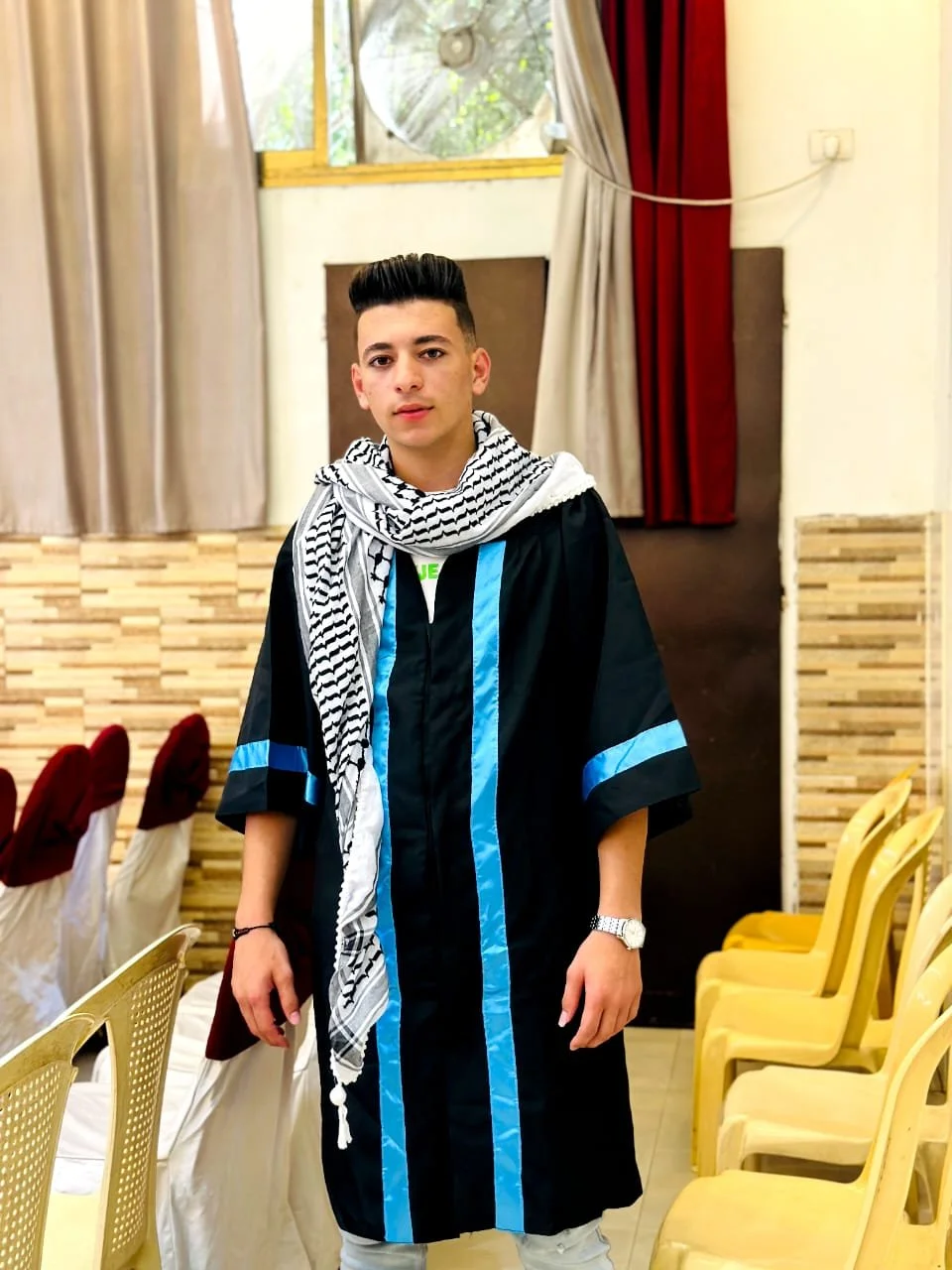 Young man in graduation attire with a black gown and blue stripes, wearing a keffiyeh scarf, standing in a decorated room with chairs and curtains.