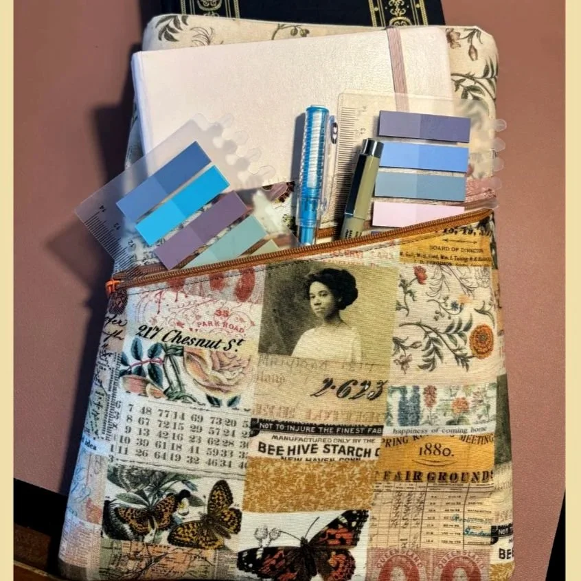 A fabric book sleeve with a zippered pocket with collage-style prints including a woman, butterflies, postage stamps, and text, situated on a pink surface with some notebooks and sticky tabs spilling out.