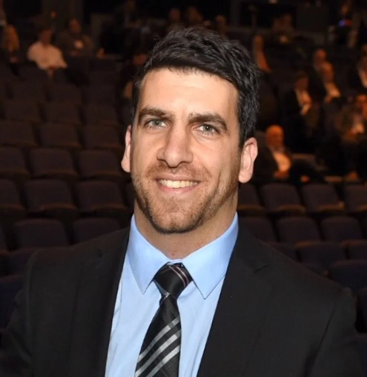 A man with dark hair and a beard, wearing a suit and tie, smiling in a large auditorium with several people in the background.