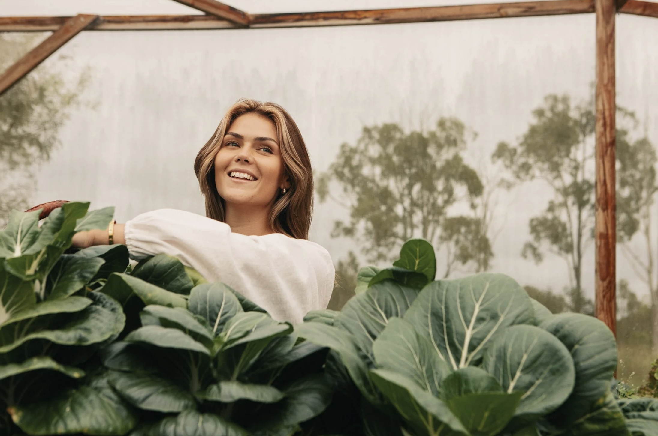 Lilly Moyes smiling in a garden, surrounded by large green leafy plants, in a greenhouse setting.