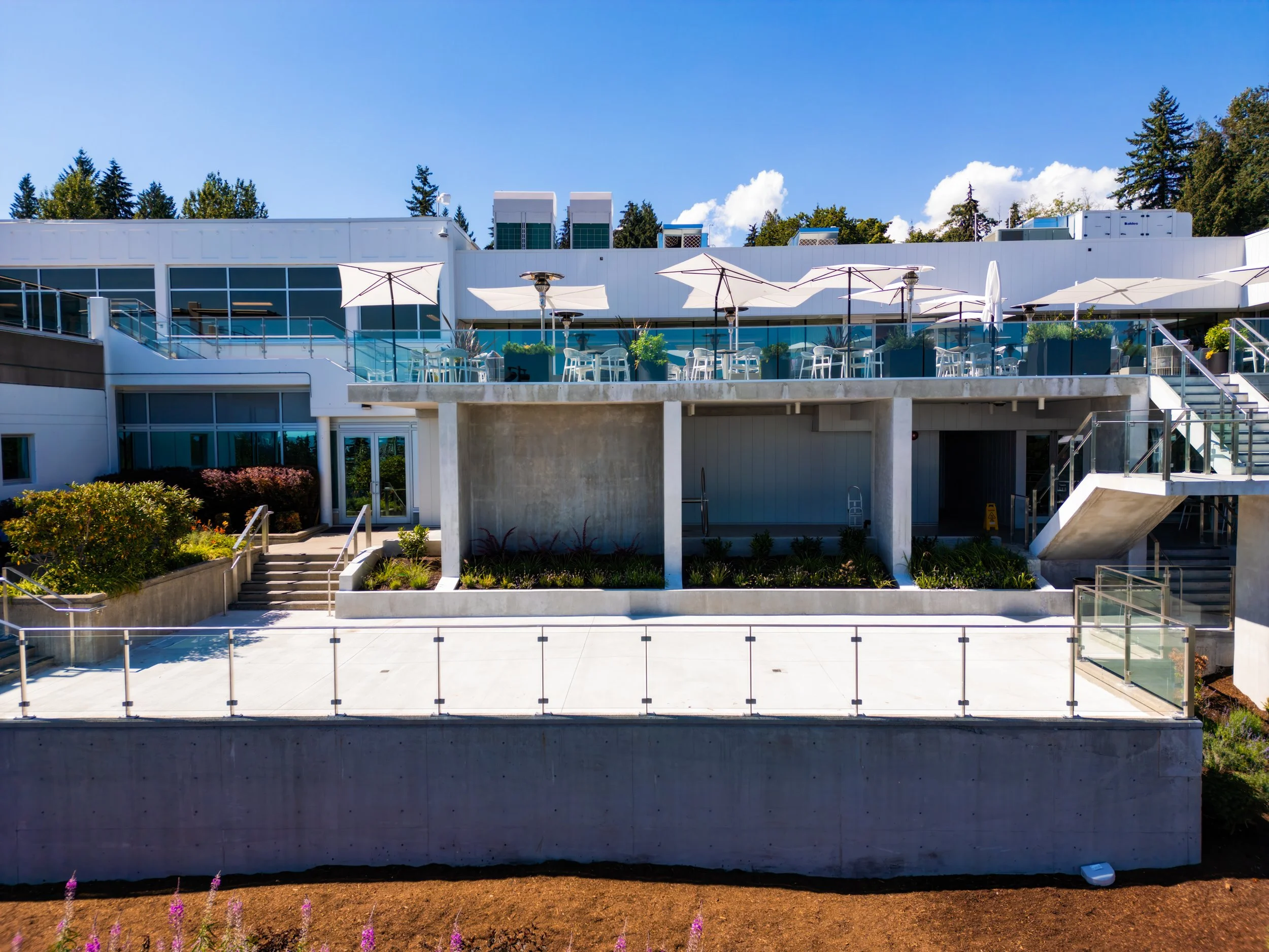 Modern multi-story building with outdoor patio area, white umbrellas, and glass railings, set against a blue sky and surrounded by trees.