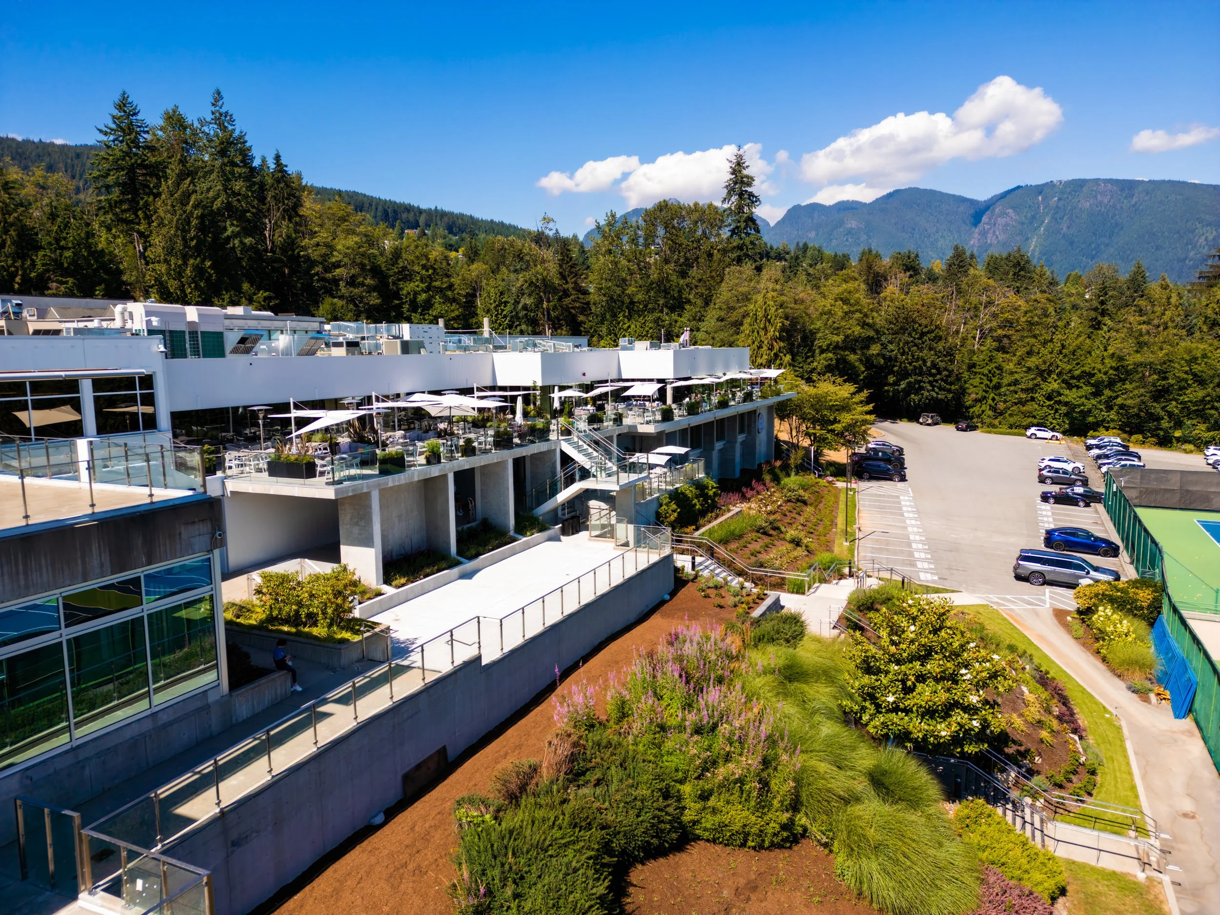 Outdoor terrace with seating and umbrellas next to a parking lot with cars, surrounded by landscaped bushes and trees, with mountain and forest background under a partly cloudy sky.