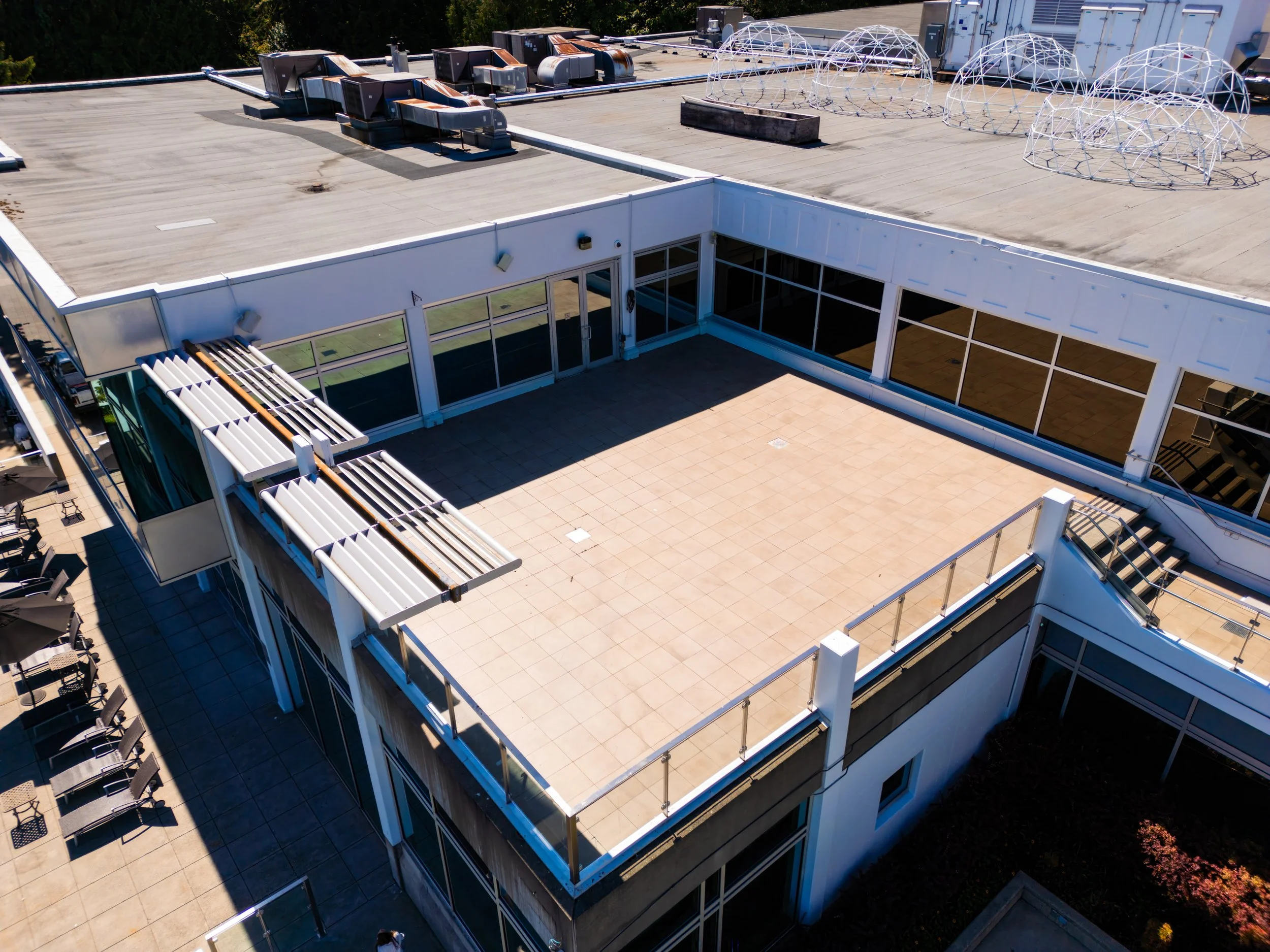 A rooftop terrace with beige tile flooring, glass railing, and rooftop structures, part of a commercial building with another rooftop area featuring vents and geodesic dome shapes.