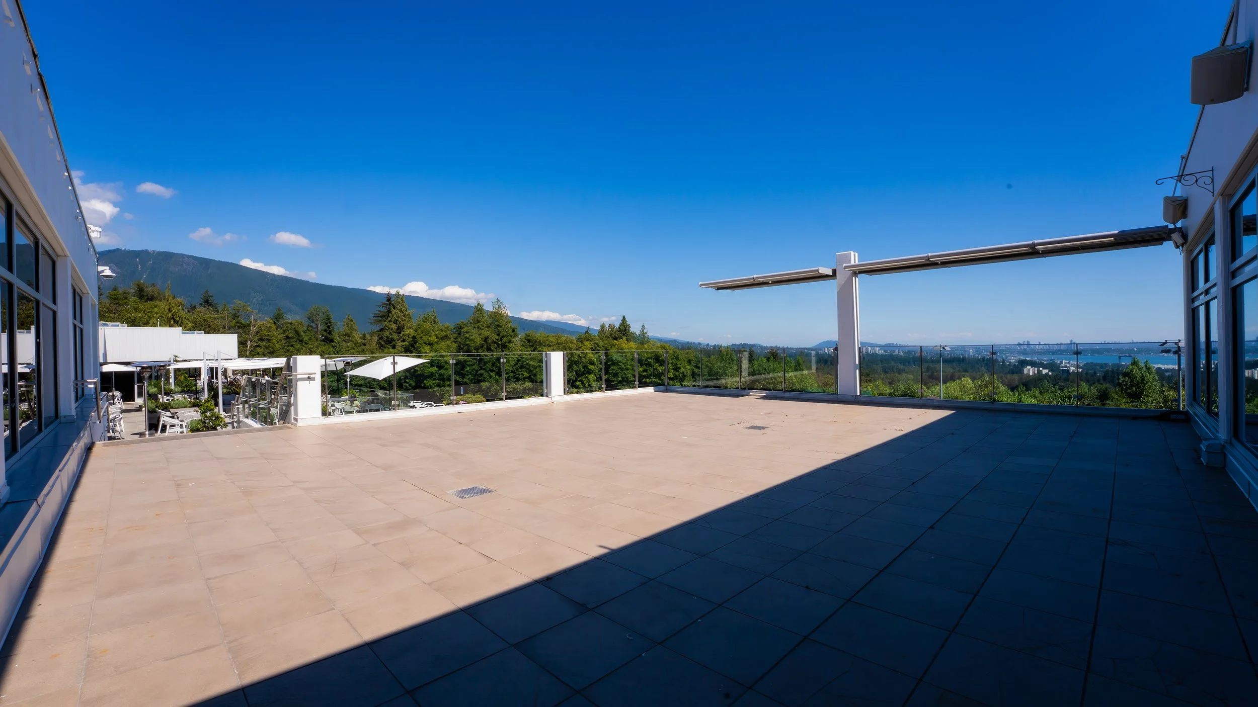 Large empty rooftop terrace with glass railing, overlooking a city, mountains, and water, under blue sky.
