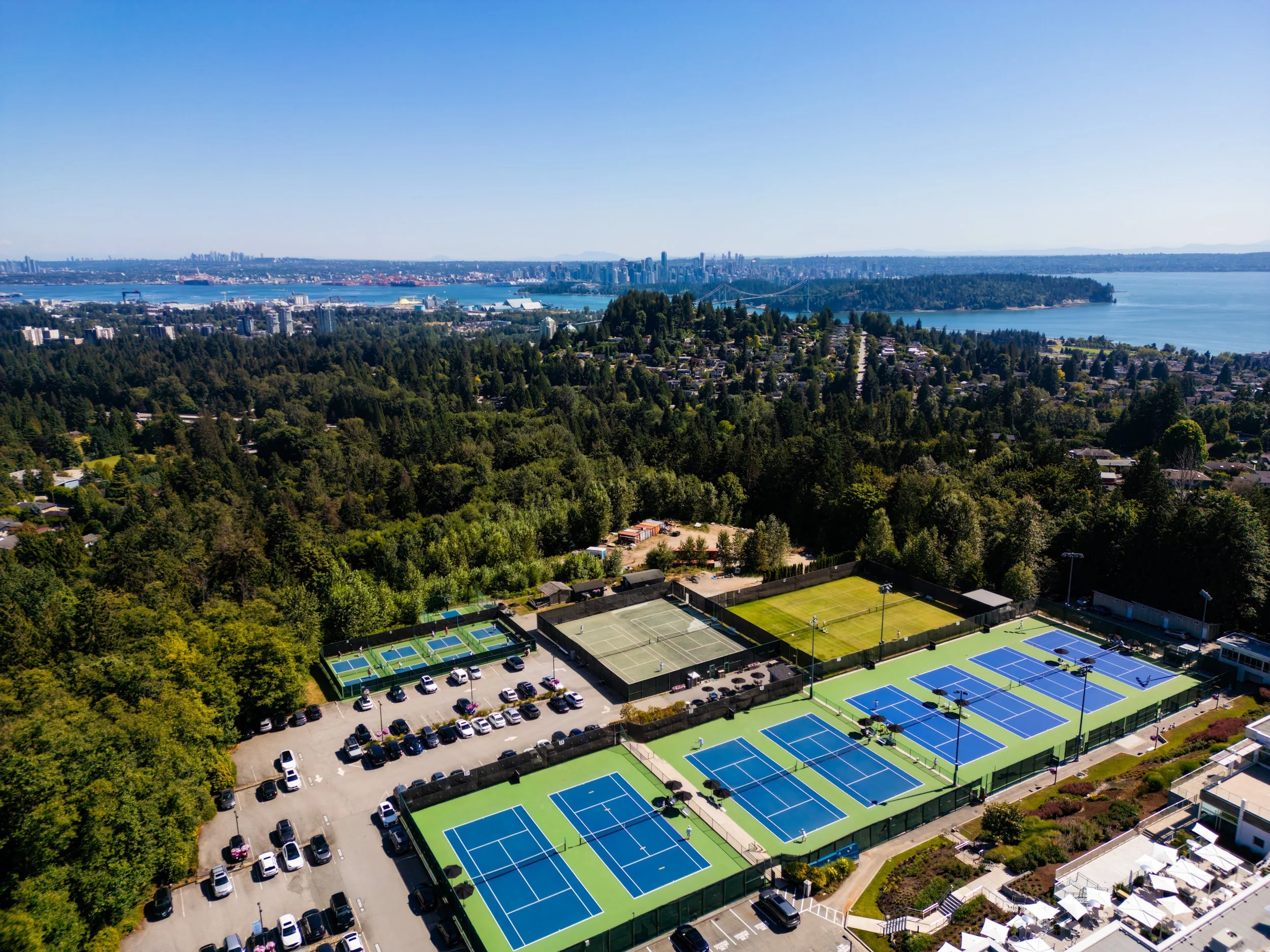 Aerial view of a tennis facility with multiple courts and a parking lot surrounded by greenery, overlooking a city skyline and water.