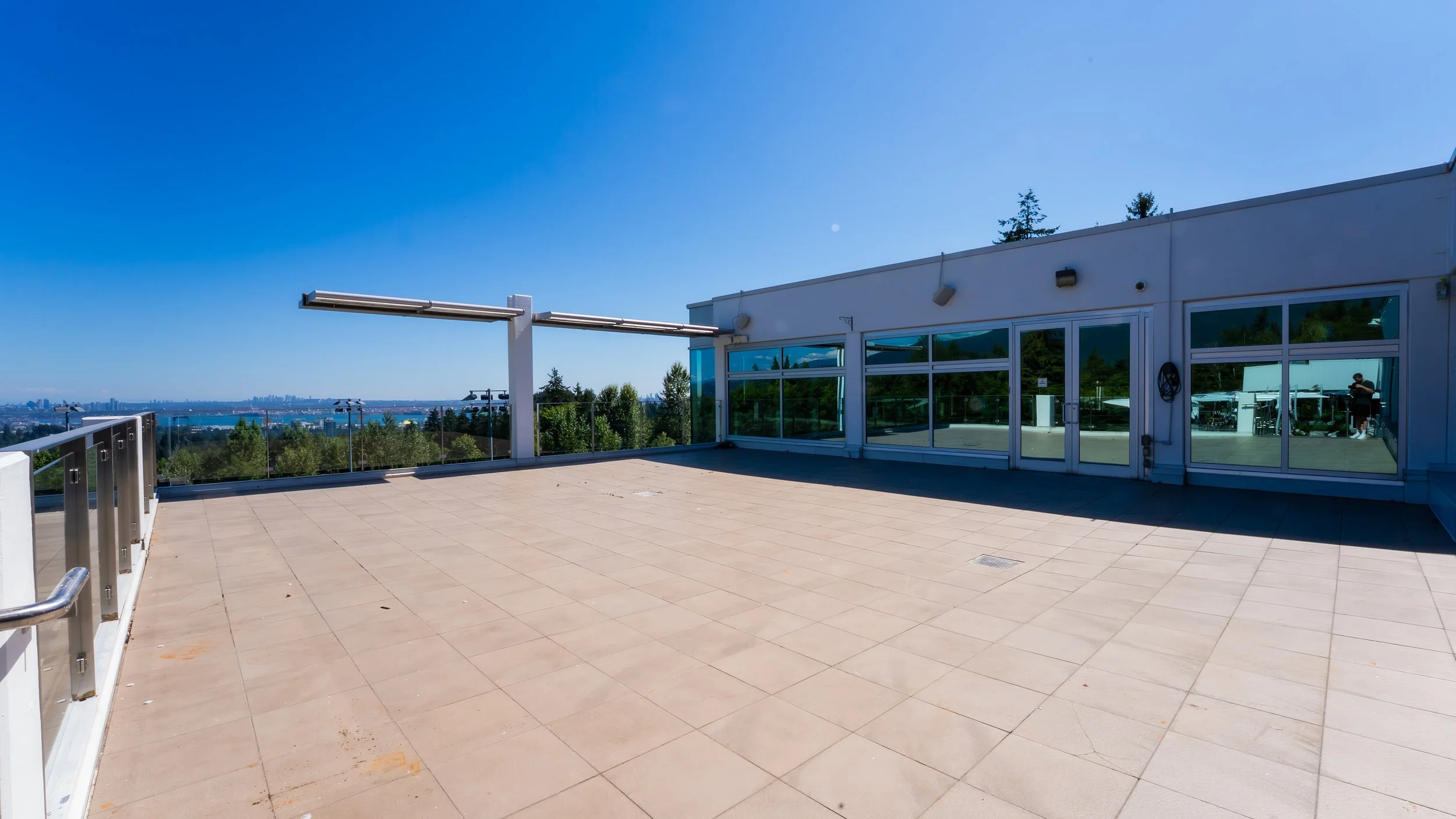 Empty rooftop terrace with glass railings, view of city skyline and water in the distance, clear blue sky, and a building with large windows.
