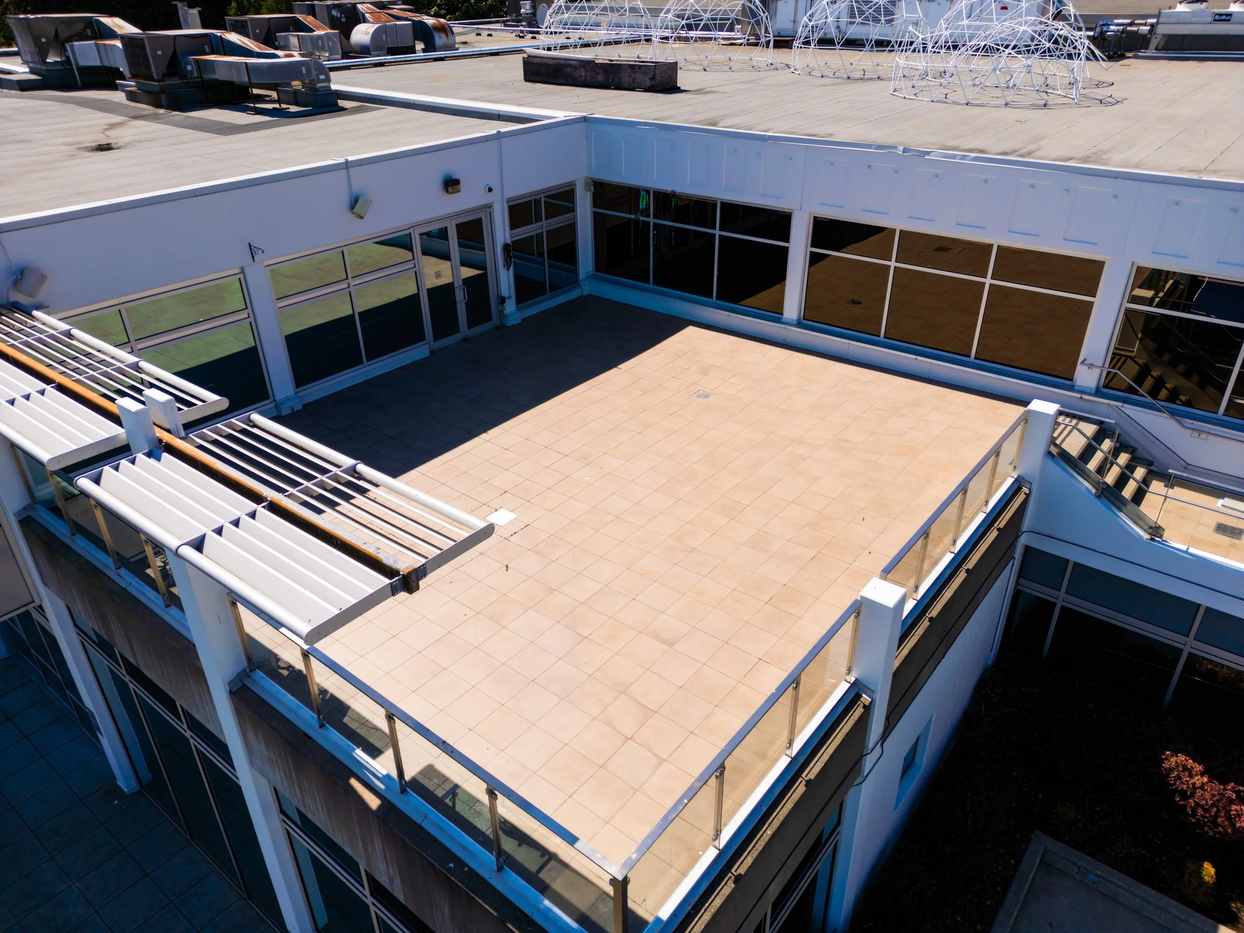 Large rooftop patio with beige tiles, glass railings, and surrounding white building structure. Some rooftop features include air conditioning units, ventilation systems, and climbing structures or geodesic domes.