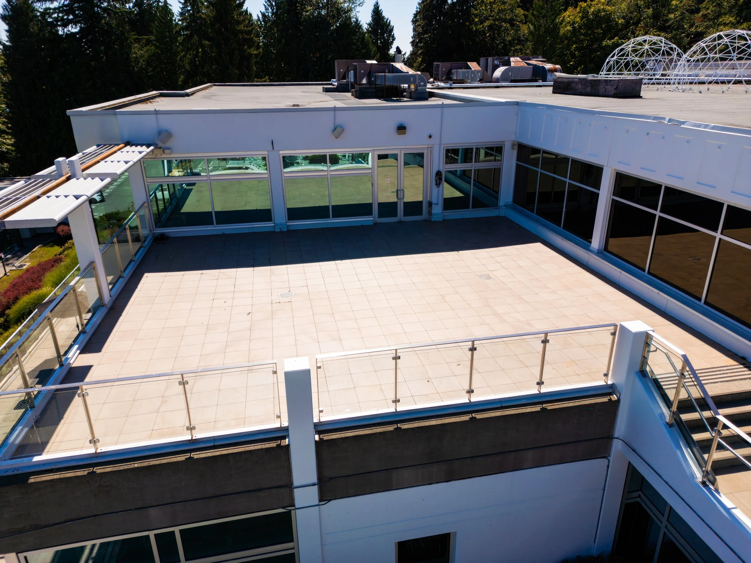 Empty rooftop terrace with glass railing, large glass windows, and a small set of stairs on the right, with surrounding trees and a blue sky.