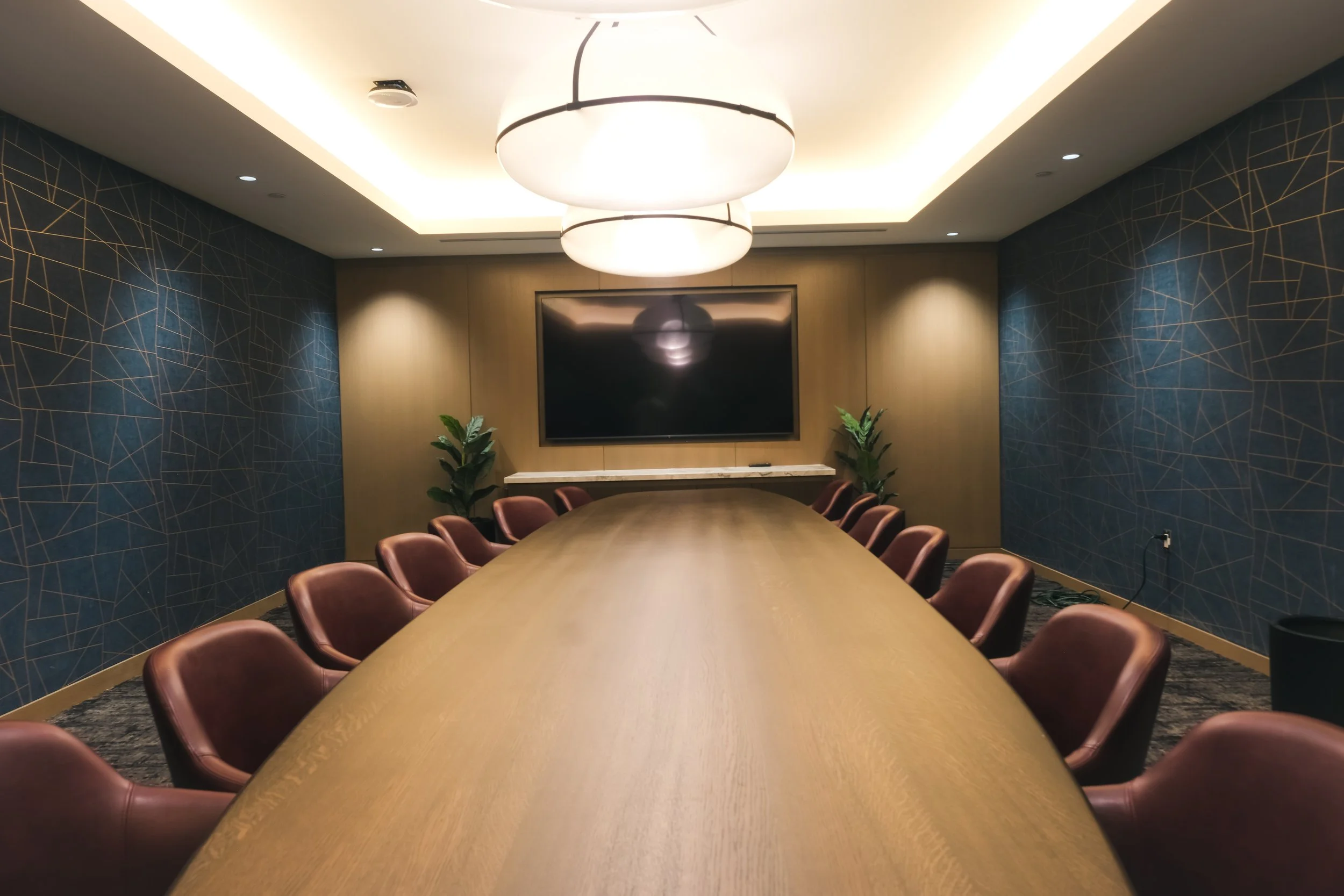 Modern conference room with an oval wooden table, leather chairs, large TV screen, ceiling lights, and decorative wall design.