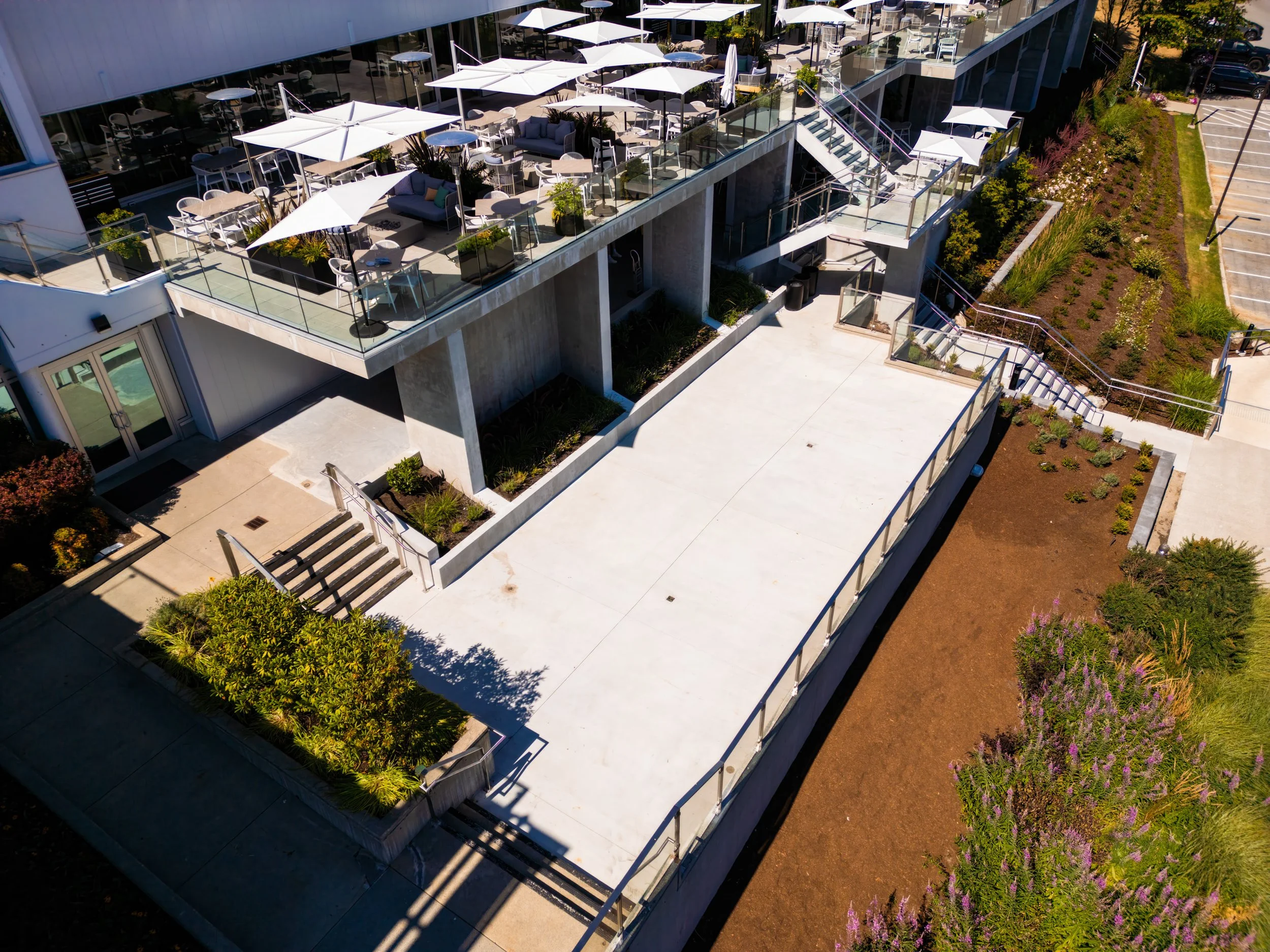 Aerial view of a modern outdoor terrace with tables, chairs, and umbrellas, adjacent to a landscaped area with plants and a parking lot.