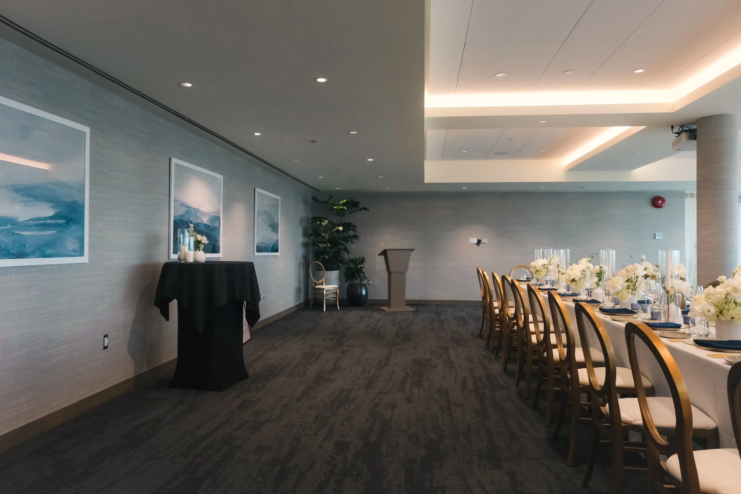 Empty banquet room with a long table set with white flowers, glassware, and napkins, a podium at the front, and wall art depicting blue landscapes.