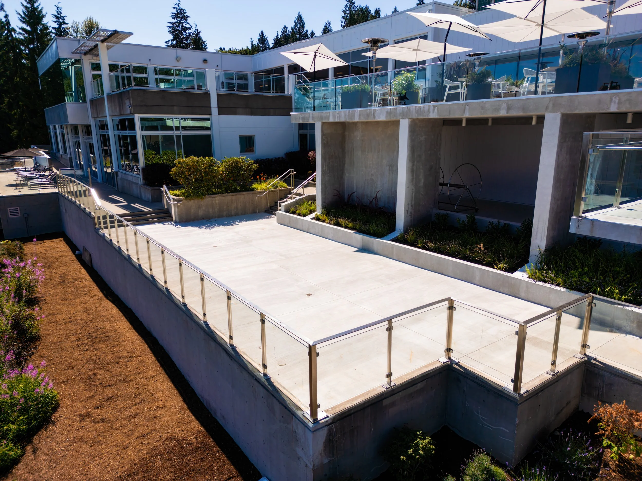 Modern building with glass windows, outdoor terrace with umbrellas, and a concrete walkway with plants and a glass railing.