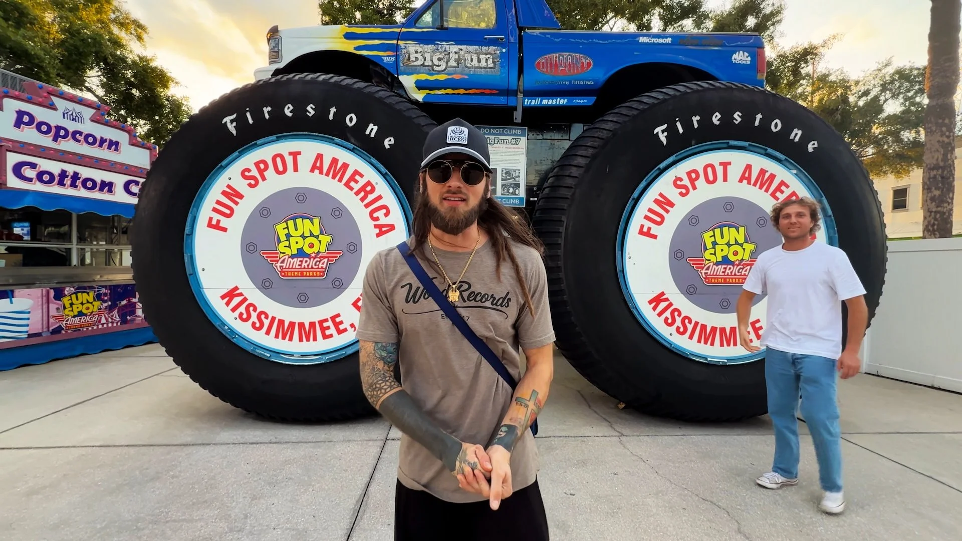 Two young men stand in front of giant tires at Fun Spot America amusement park. One man has long hair, sunglasses, a cap, and tattoos on his arms. The other man has curly hair and is wearing a white t-shirt and jeans. In the background, there is a colorful popcorn stand.