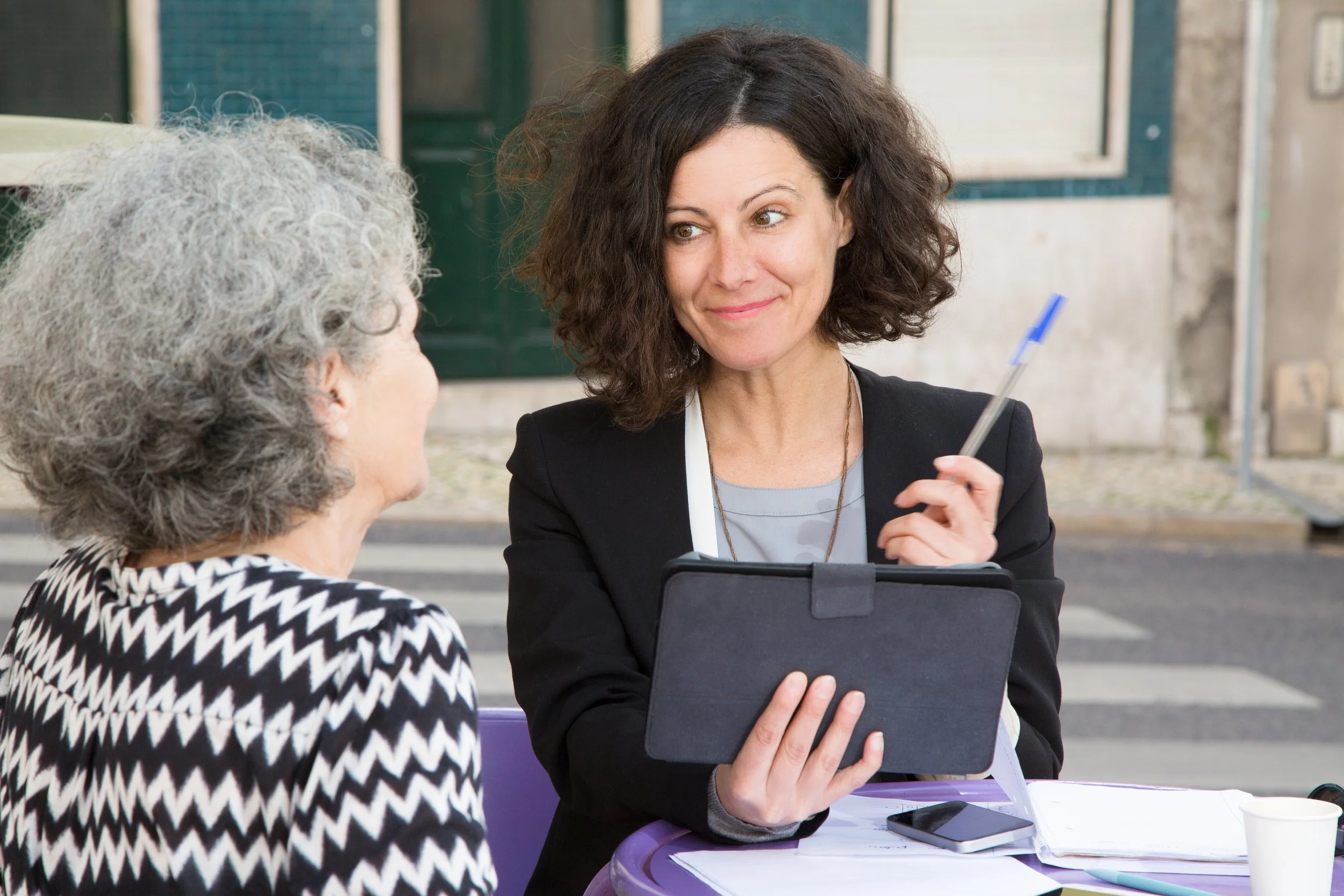 Two women sitting at a purple outdoor table, having a conversation. The woman on the right, with curly brunette hair, is holding a tablet and a pen, smiling at the older woman on the left who has gray hair and is wearing a black and white patterned top.