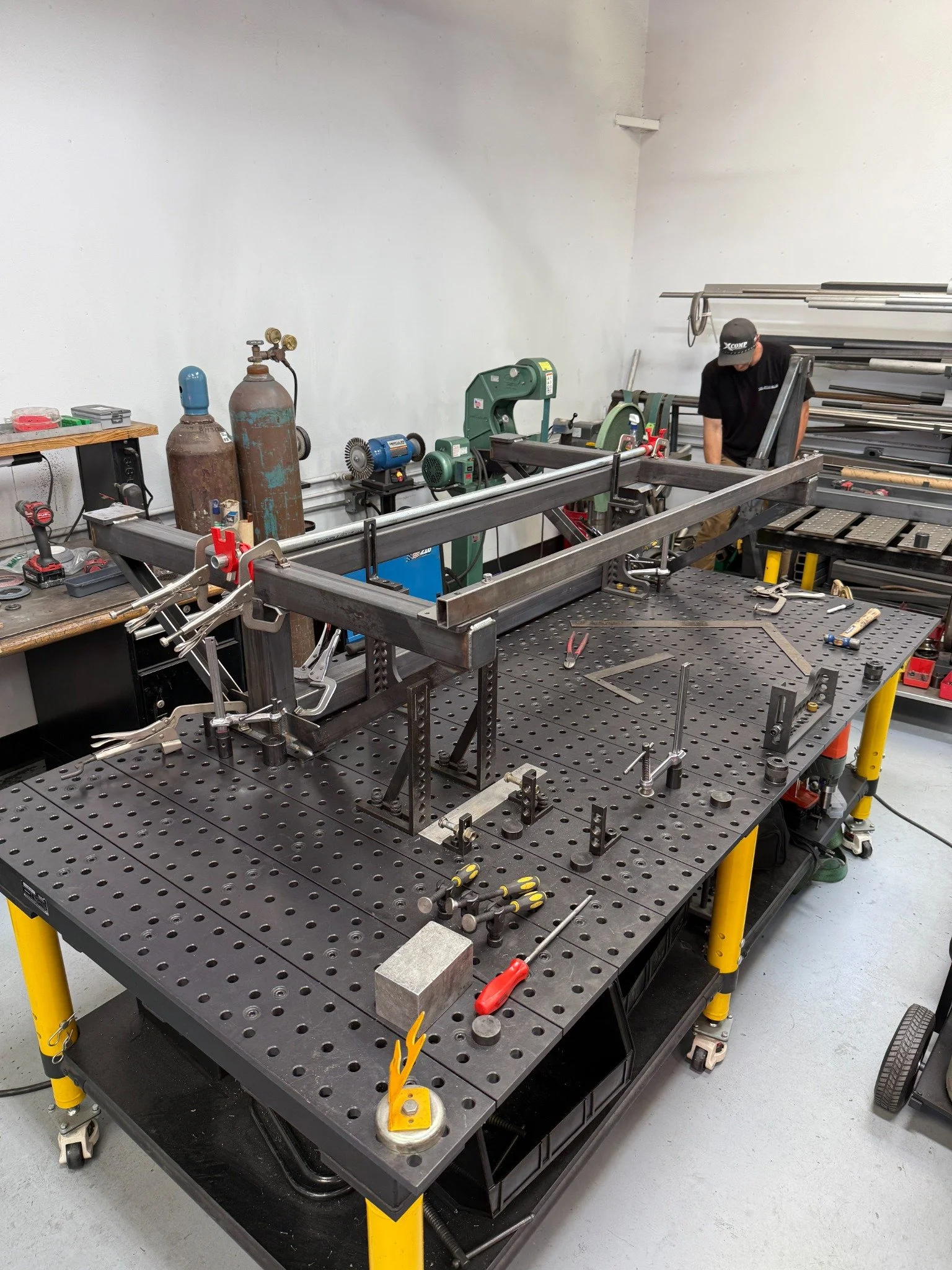 Steel tube frame clamped to a perforated fixture table in the metal fabrication shop.